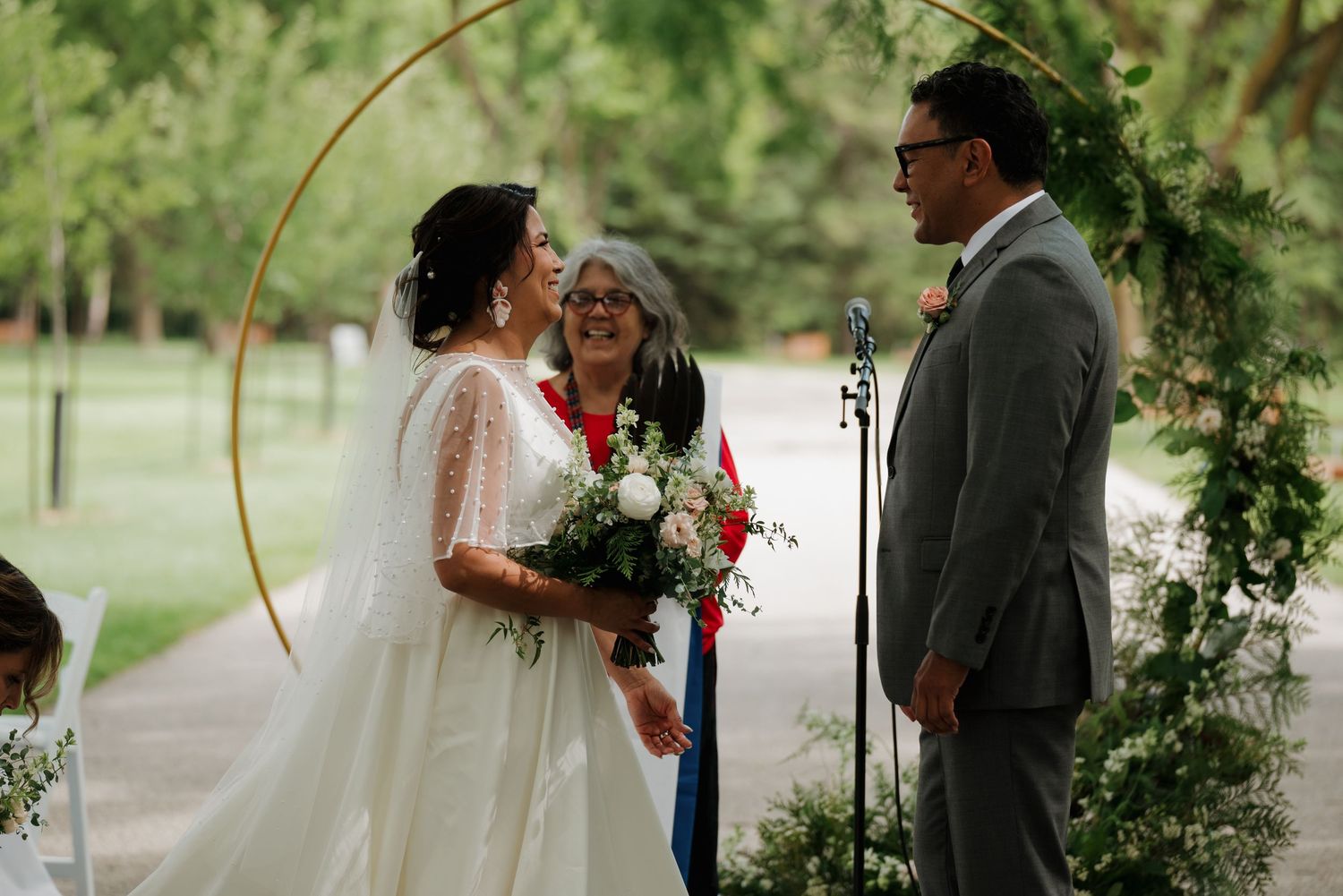 Wedding ceremony taking place under a golden arch with greenery in an outdoor garden setting.