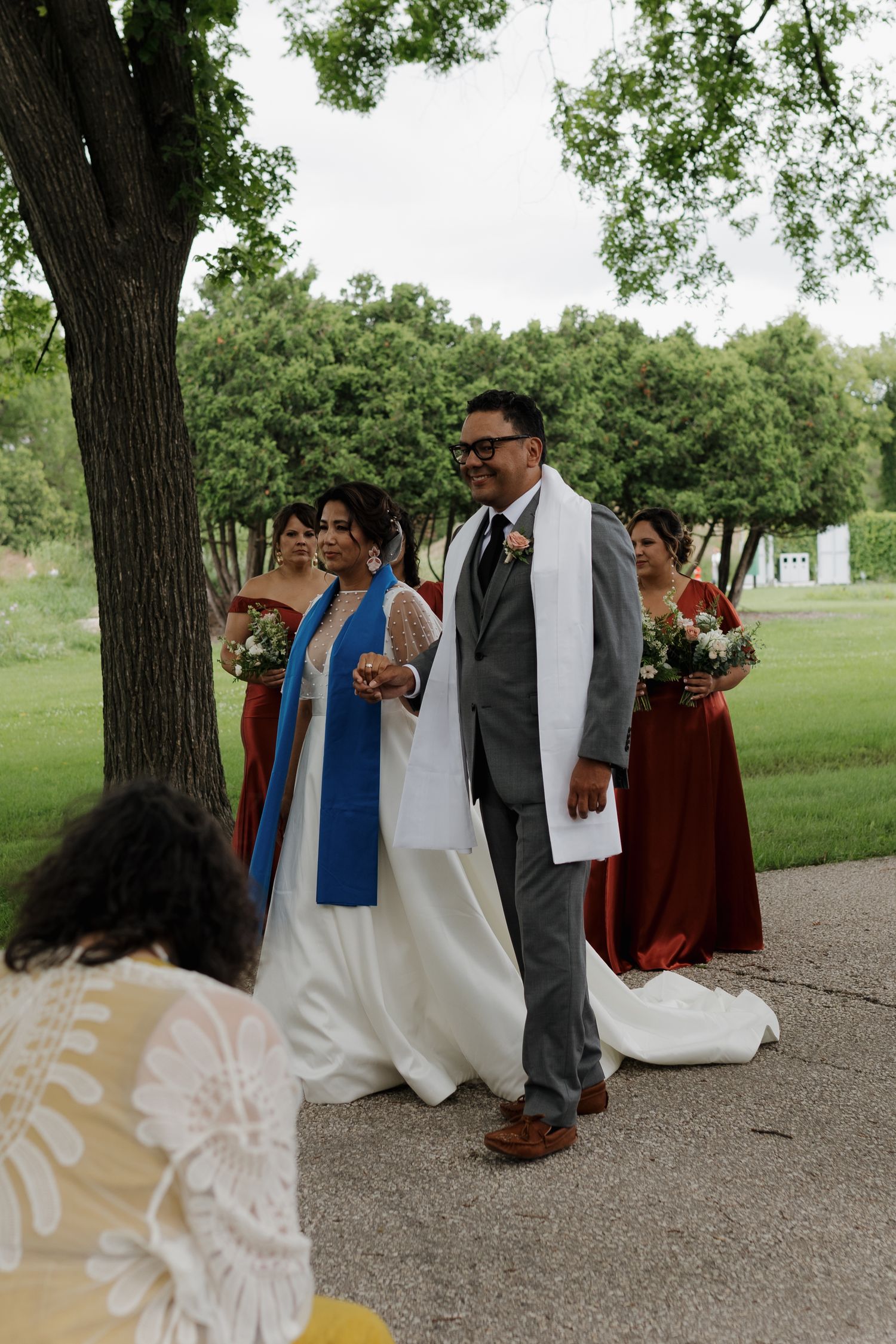 A wedding party shares a joyful moment under large trees on a gravel path during an outdoor ceremony.