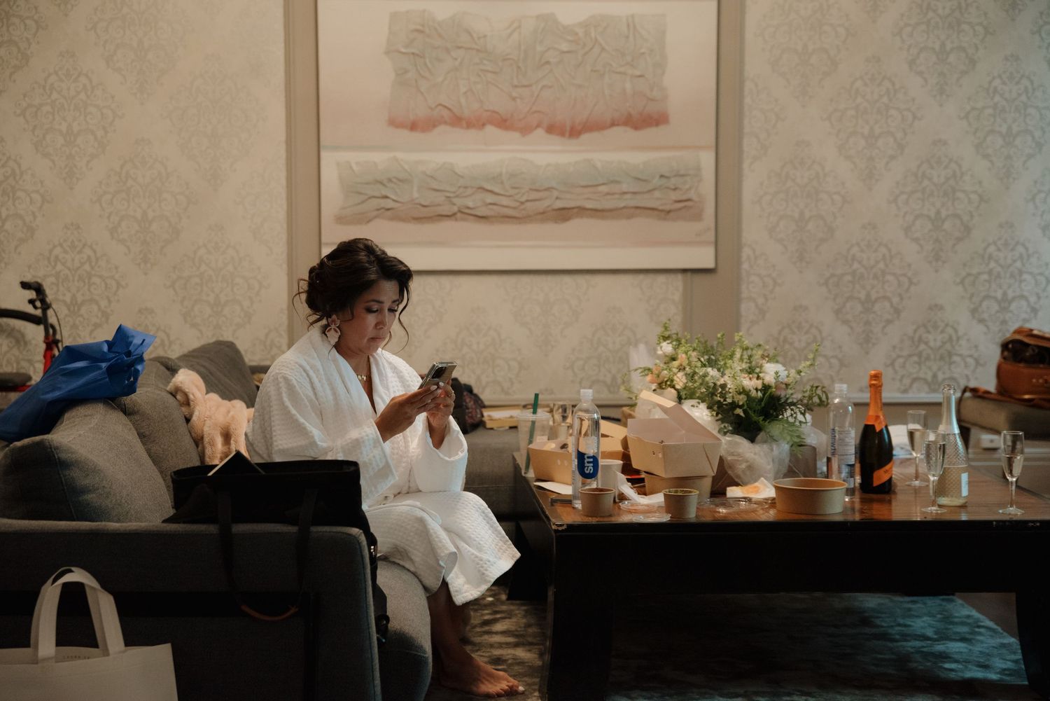 Person in white robe checking phone while surrounded by beauty products and flowers on wedding morning.