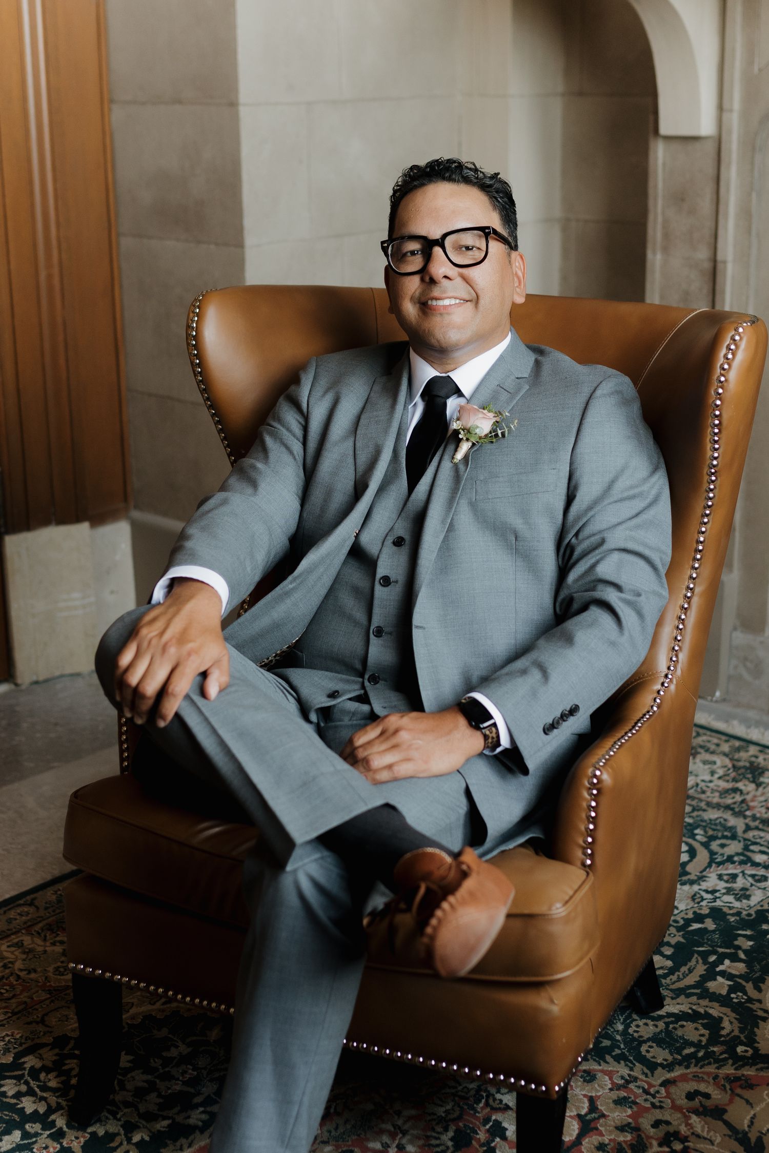Person in gray three-piece suit and glasses sitting in leather wingback chair.