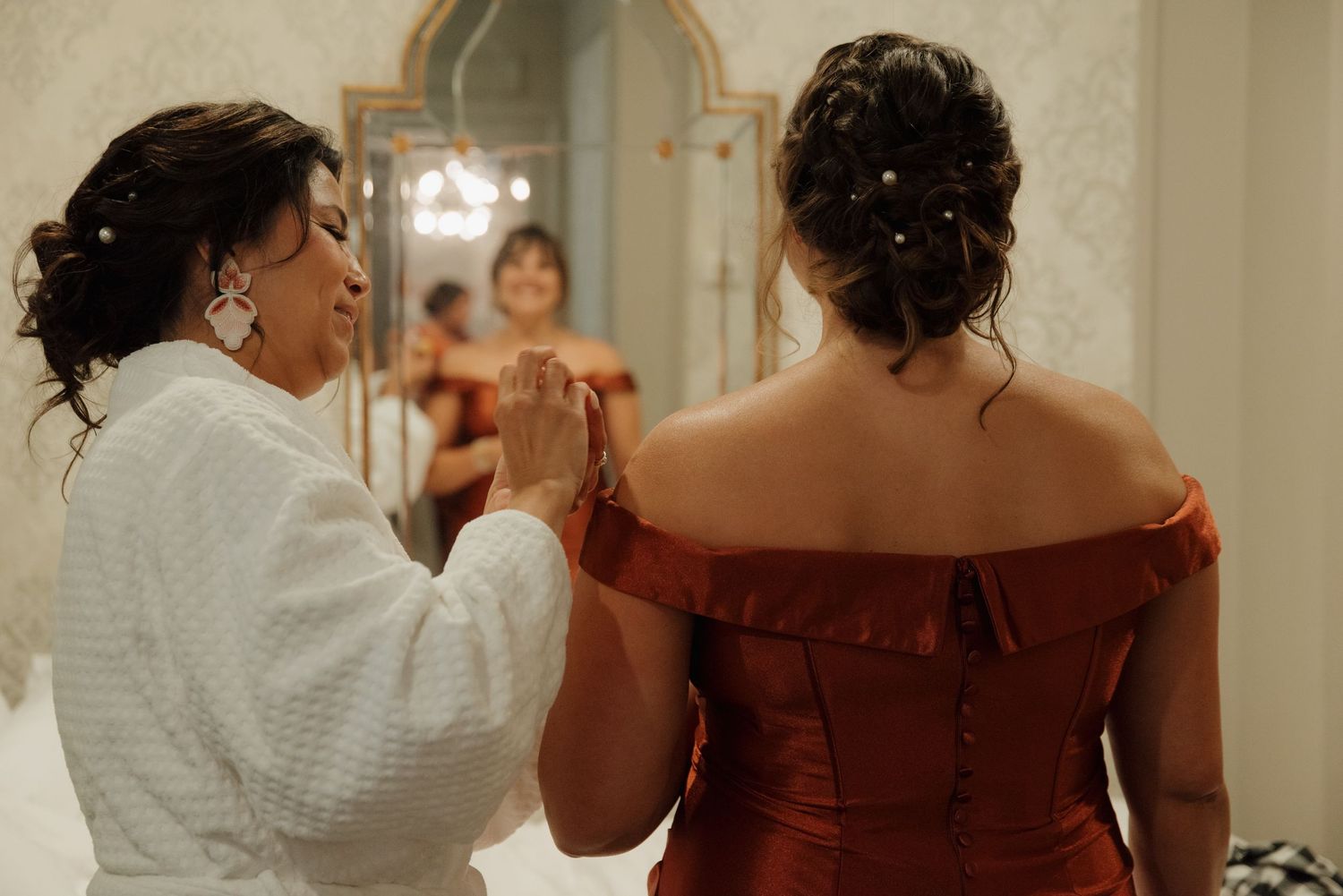 Someone helps adjust a burgundy off-shoulder dress in an elegant dressing room setting.