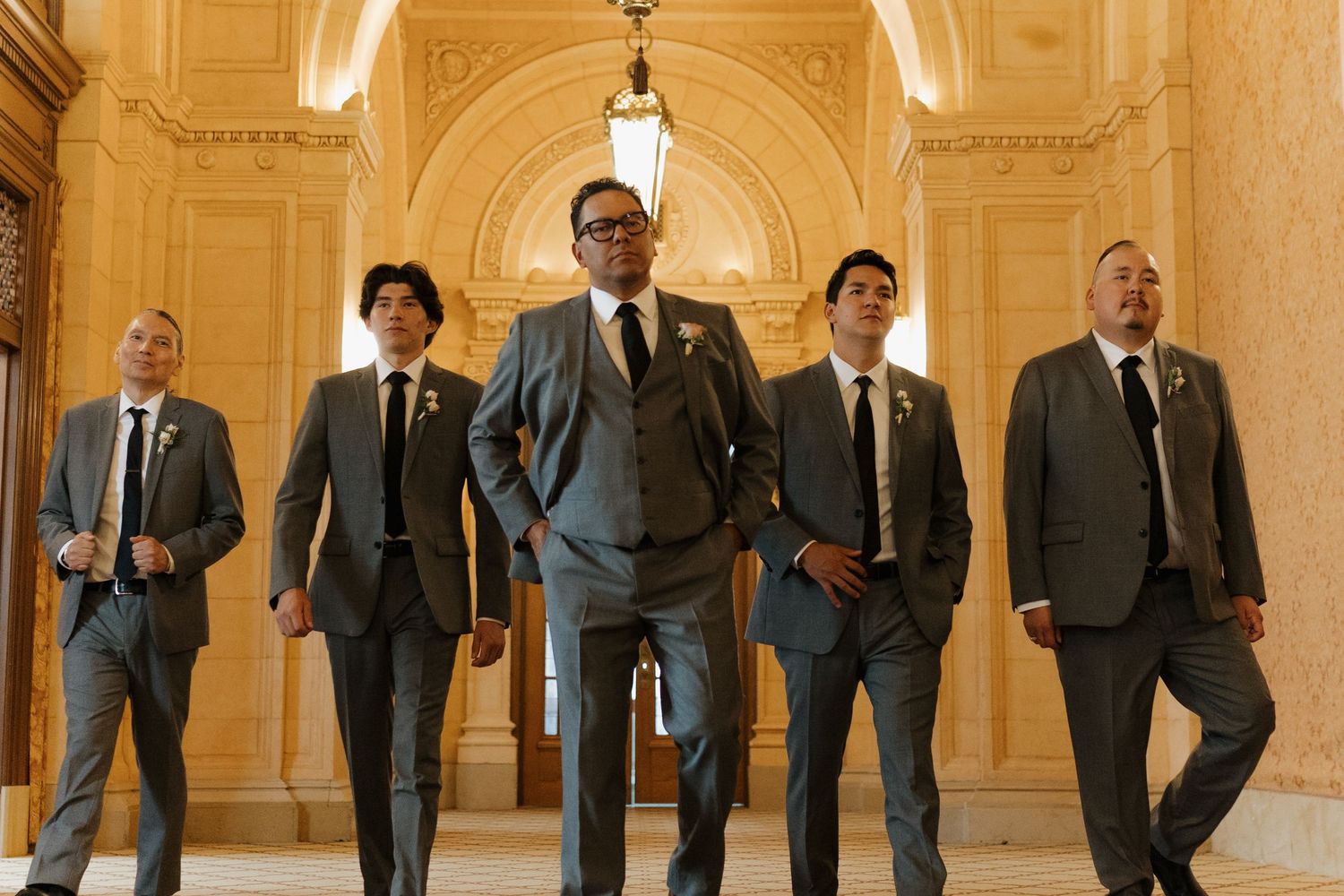 Group of groomsmen in suits posing confidently in an ornate hallway with architectural details.