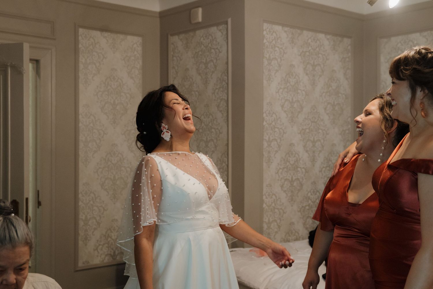 A joyful moment of shared laughter in a bridal dressing room with elegant wallpapered walls.