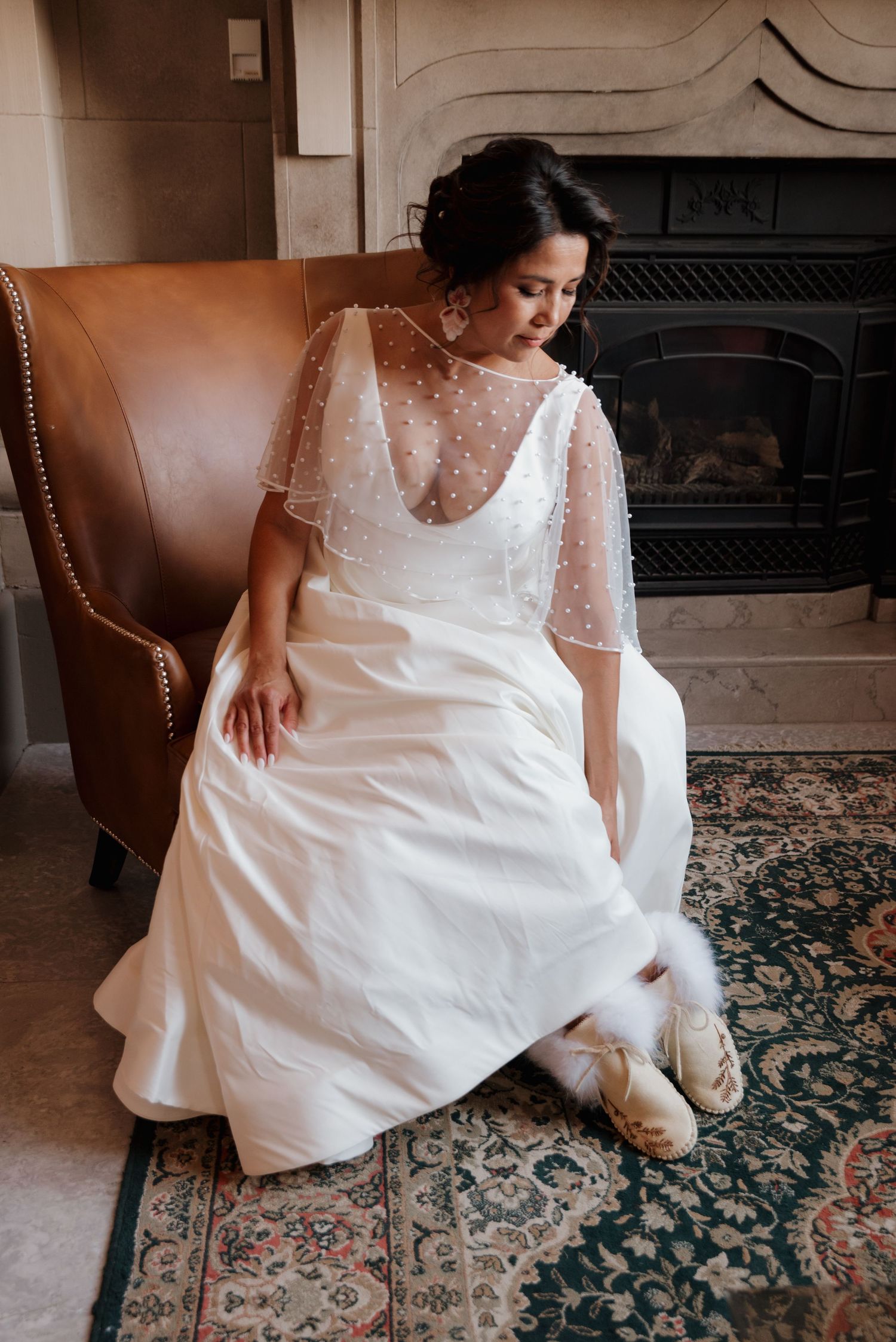 A white wedding dress with intricate details drapes elegantly across a leather chair on a patterned carpet.