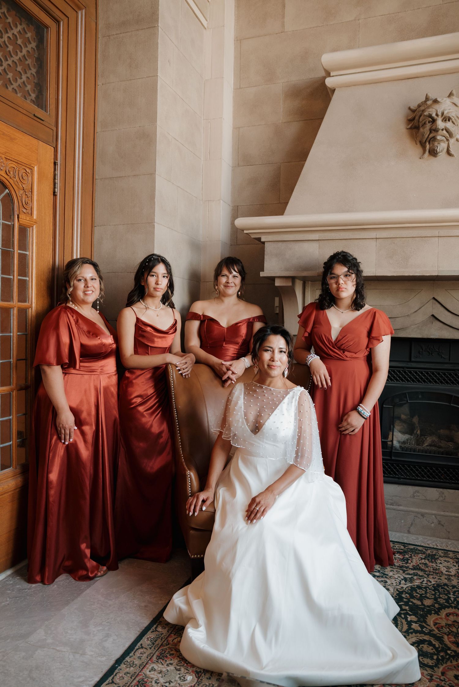 Wedding party in rust-colored bridesmaid dresses and white bridal gown posing by an elegant fireplace.