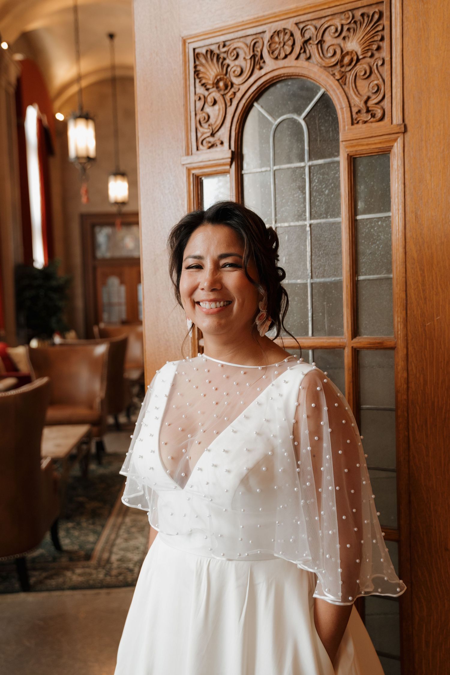 A smiling person in a white polka dot wedding dress with sheer overlay stands by an ornate wooden doorframe.
