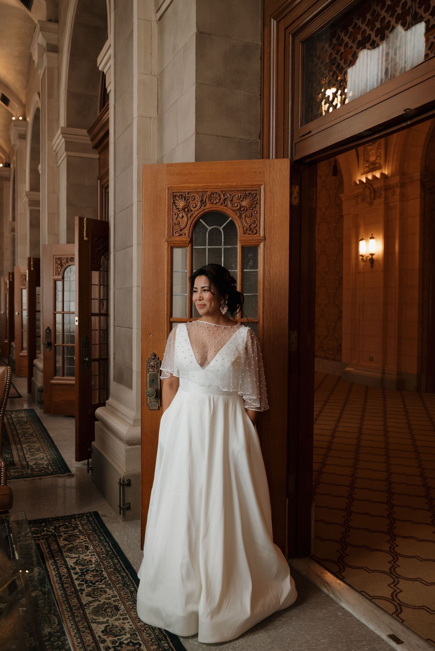 Person in elegant white wedding gown standing in ornate hallway with wooden architectural details.