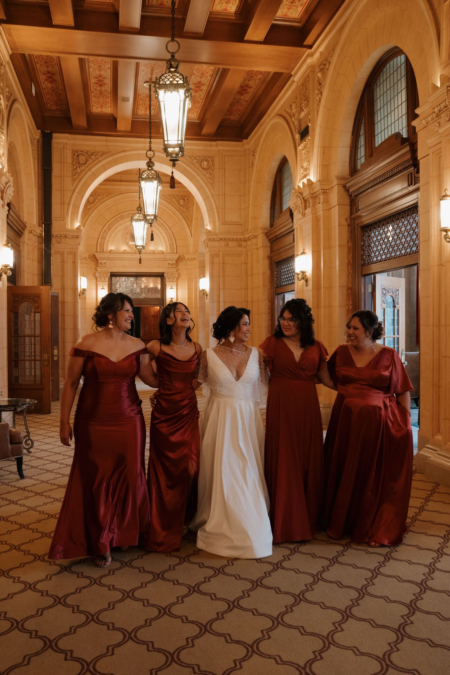 The bridal party poses in an elegant hallway with vaulted ceilings, featuring burgundy bridesmaids dresses and a white gown.
