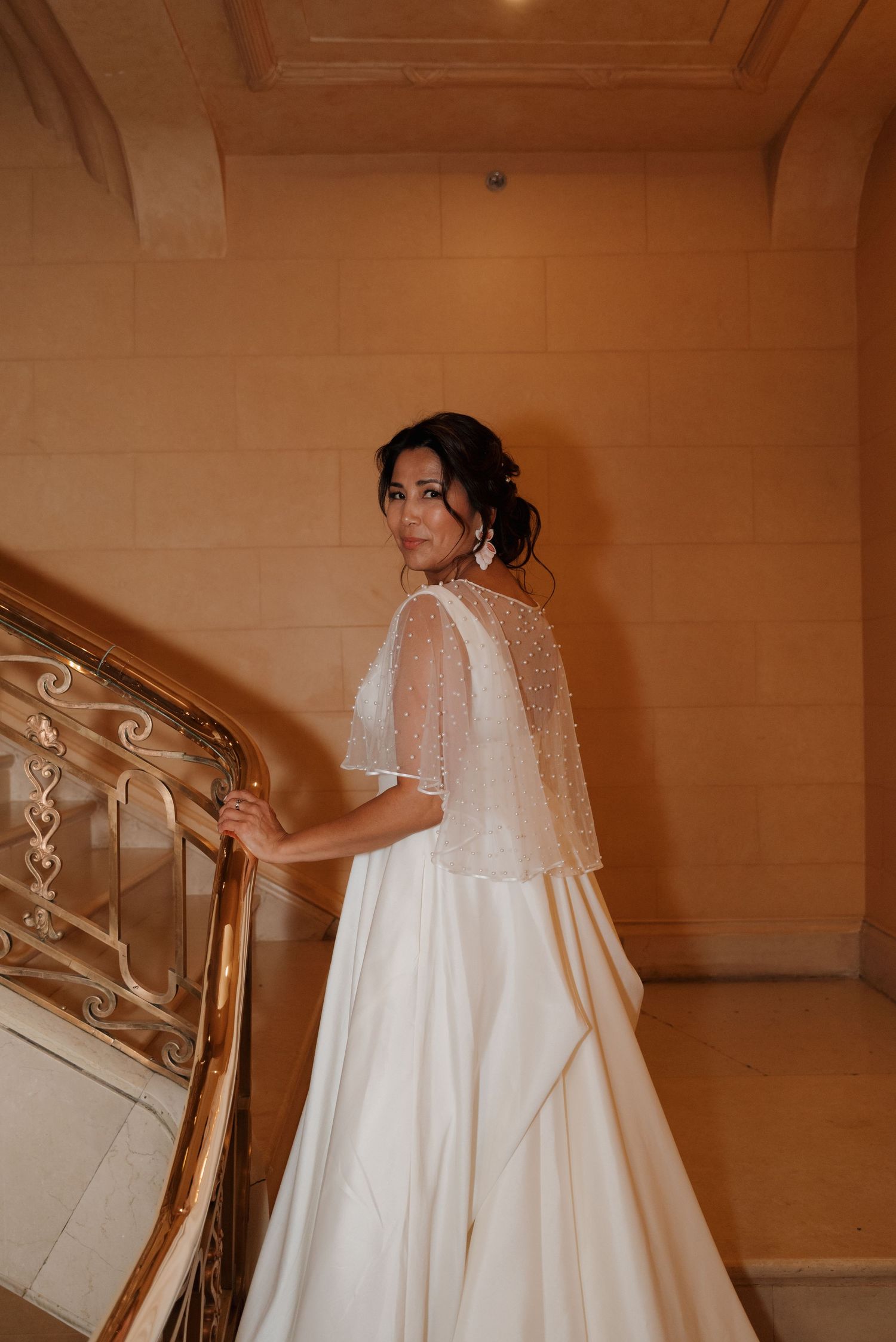 A bride in a flowing white wedding gown with sheer cape overlay poses elegantly on ornate staircase steps.
