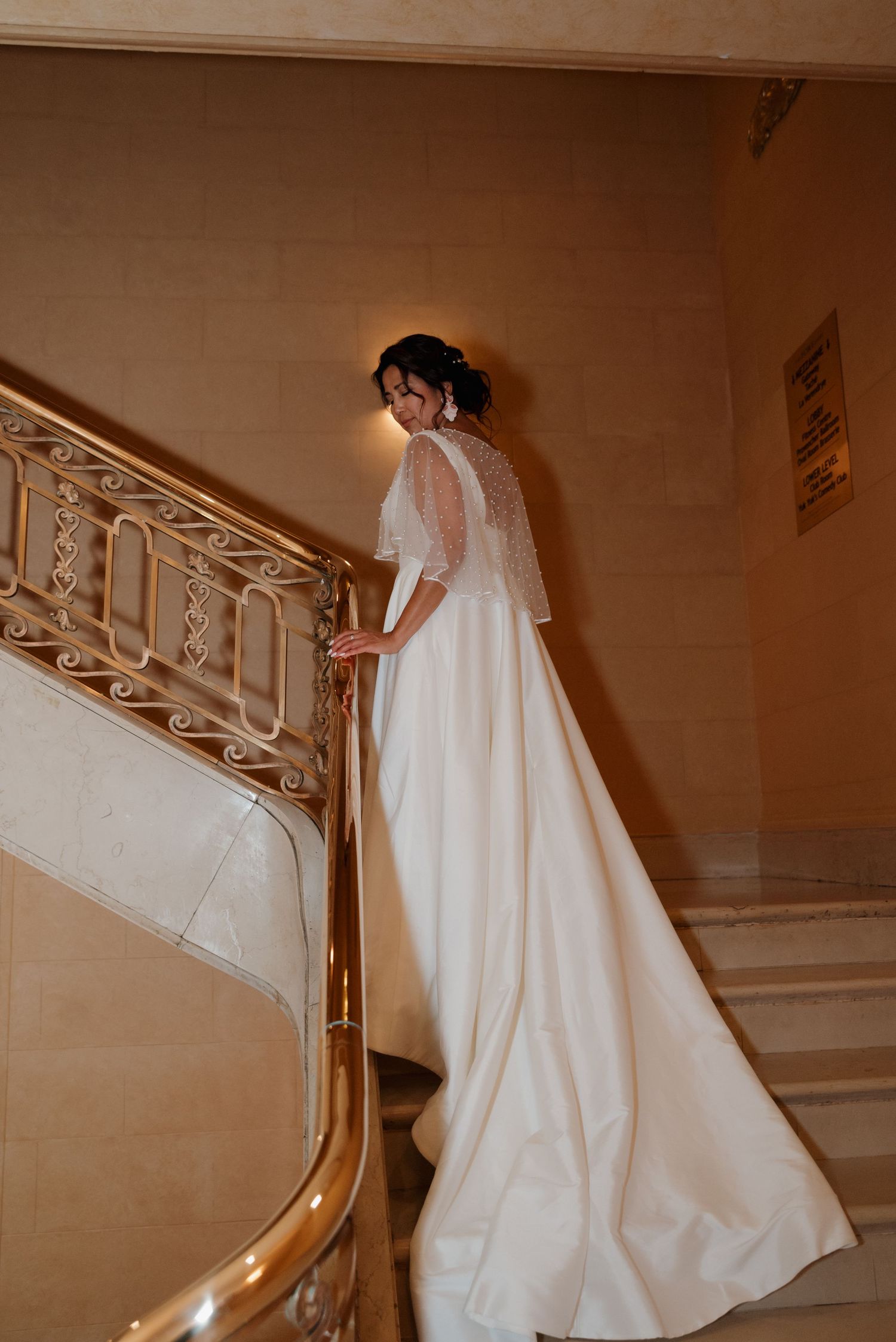 Elegant bride in flowing white dress with sheer sleeves poses on grand staircase in warm vintage lighting.
