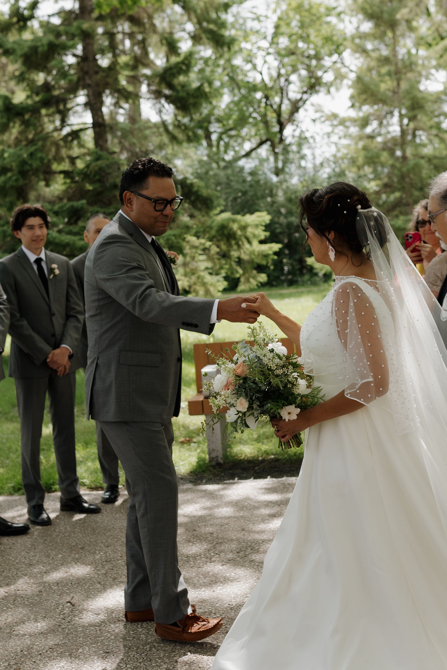 Wedding party interacting outdoors on gravel path surrounded by trees.