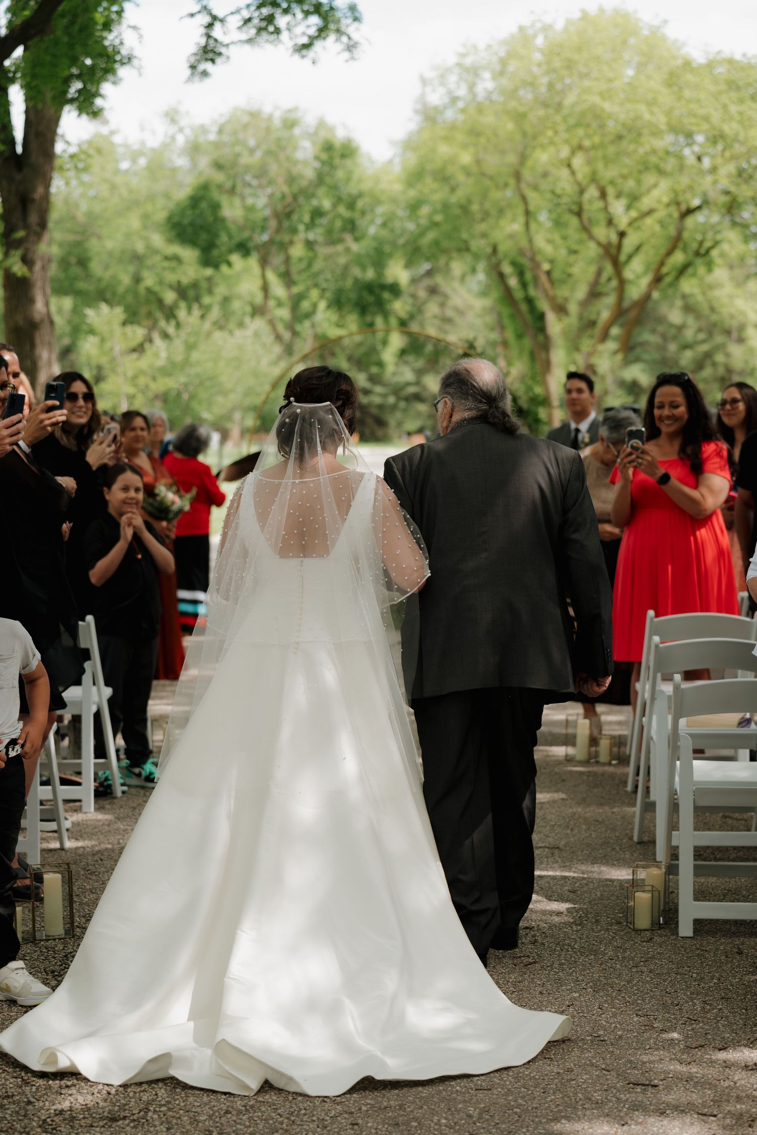 Wedding scene with bride in flowing white gown and veil walking down aisle as guests take photos during outdoor ceremony.