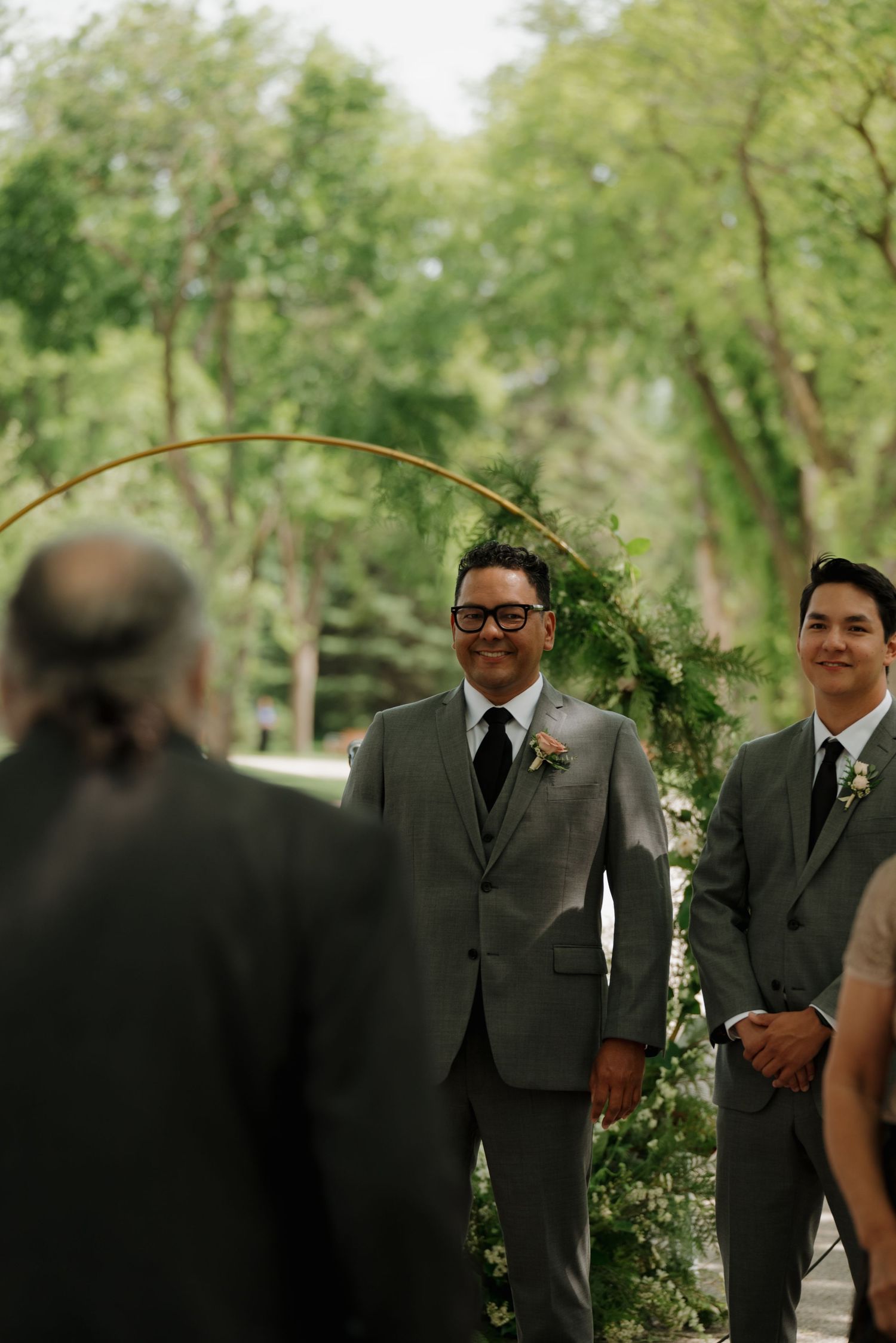 A wedding ceremony in a lush outdoor garden setting with groomsmen standing beneath a floral arch.