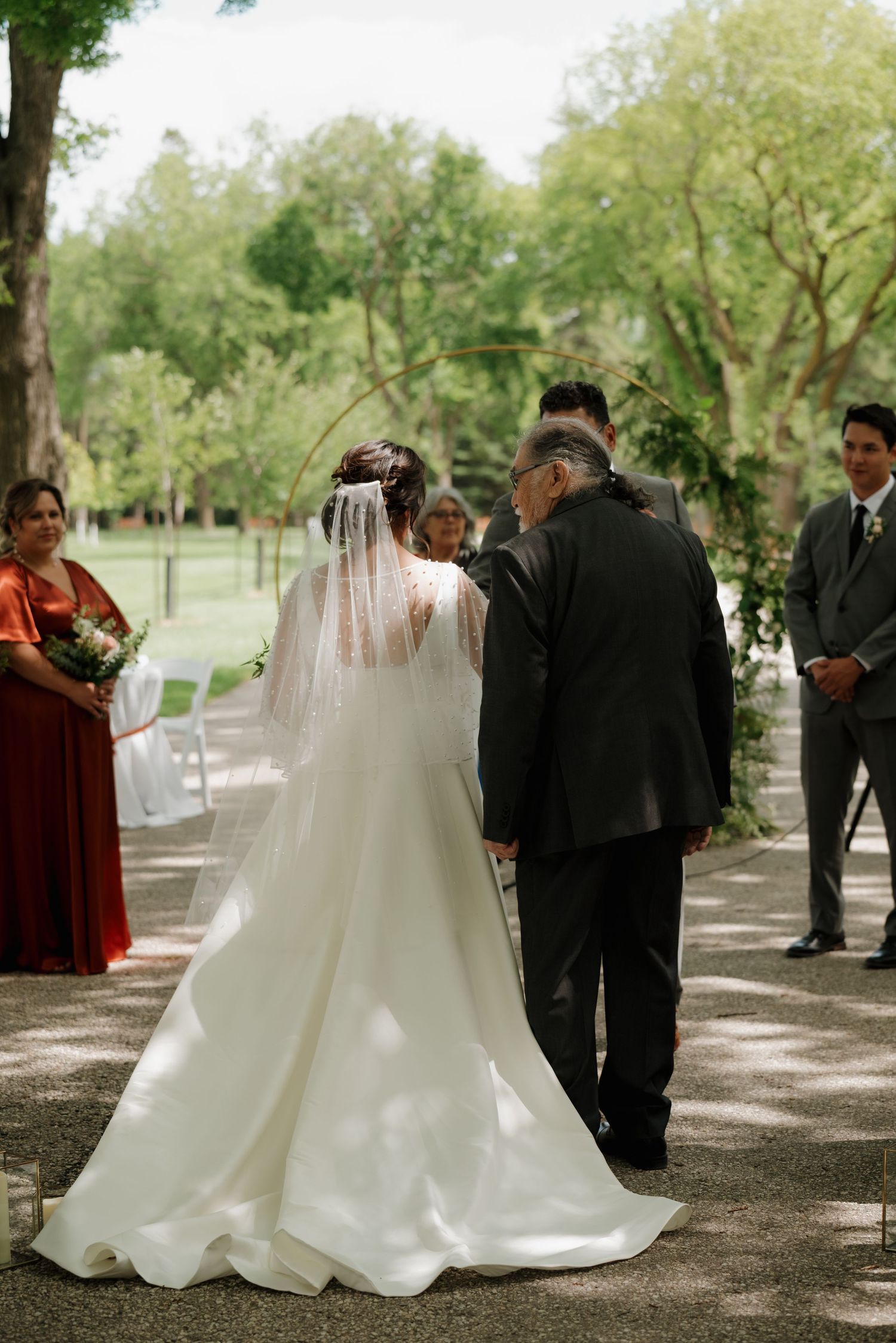 A serene outdoor wedding ceremony with a bride in a white gown and long veil walking down a tree-lined aisle.