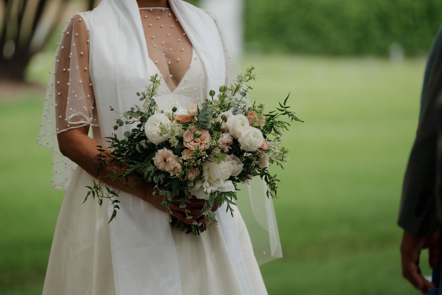 A bridal bouquet of white and blush flowers with greenery is held against a white wedding dress.