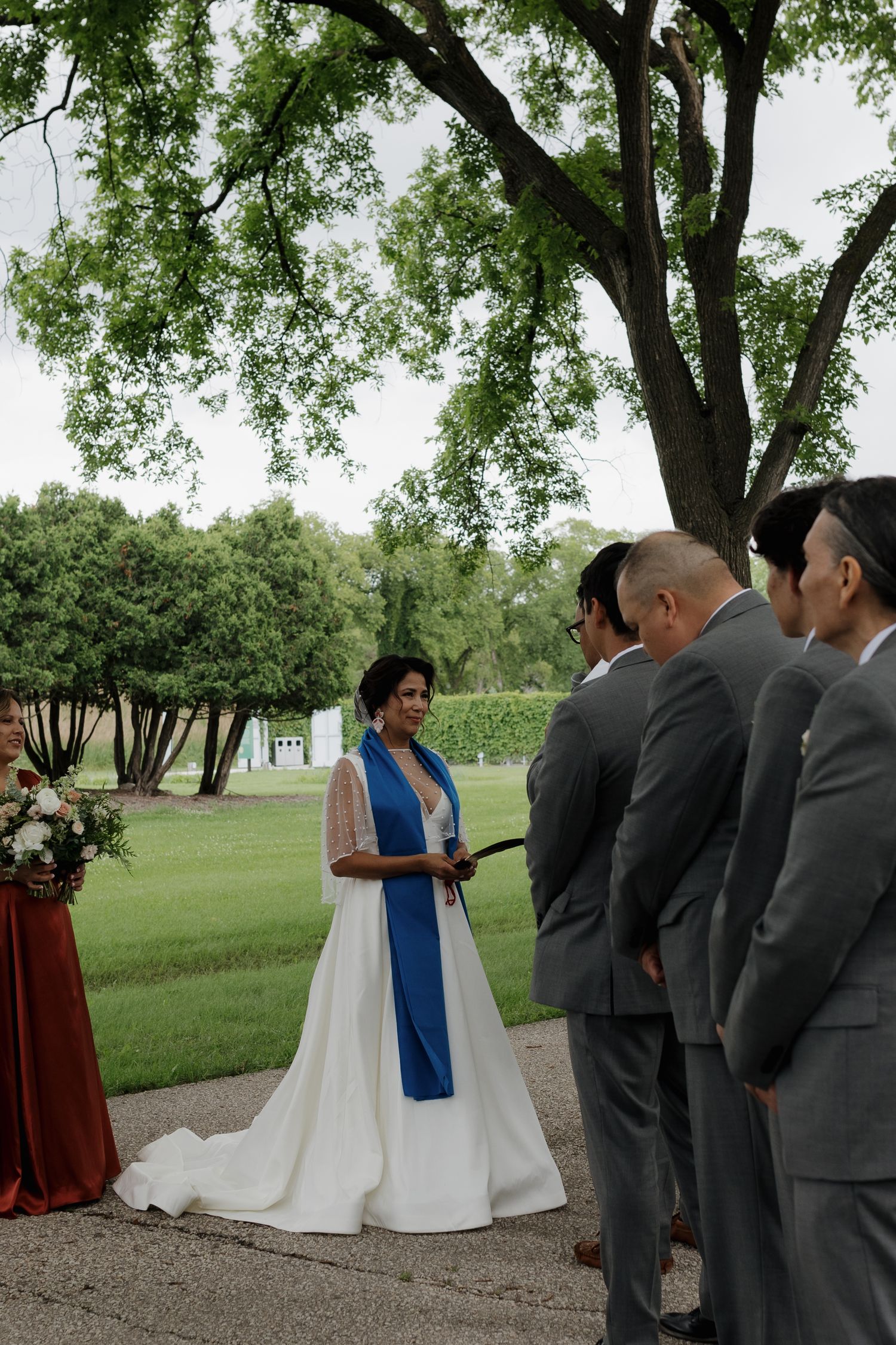 An outdoor wedding ceremony takes place under lush green trees with attendees in formal gray suits.