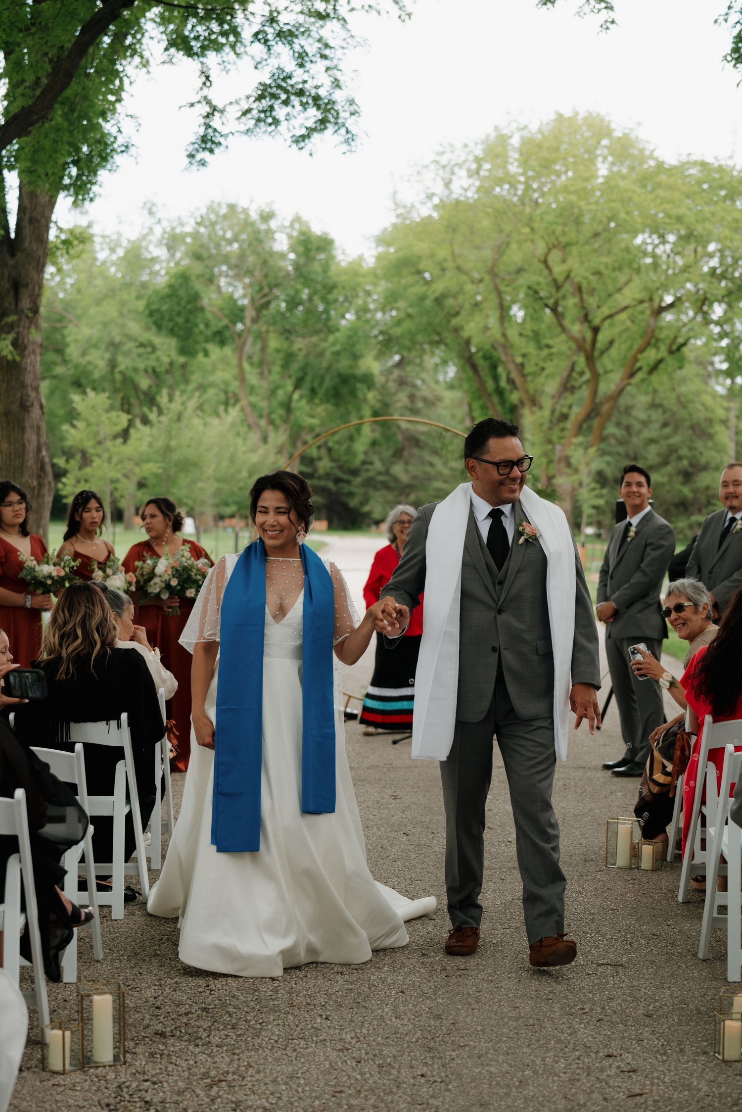 Wedding party lined up outdoors with white chairs and guests in formal attire.