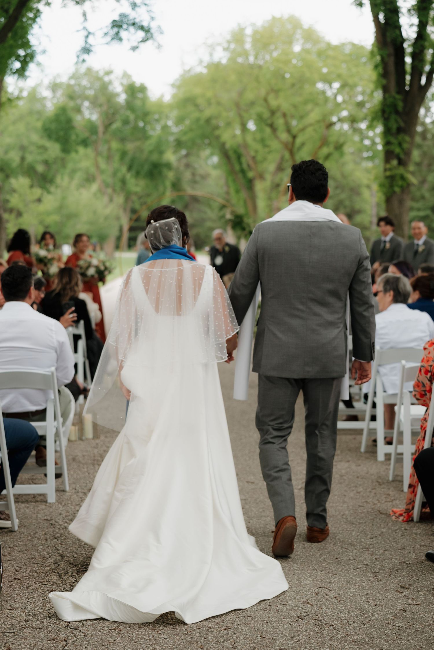 A couple walking down the aisle at an outdoor wedding ceremony surrounded by guests seated on white chairs.