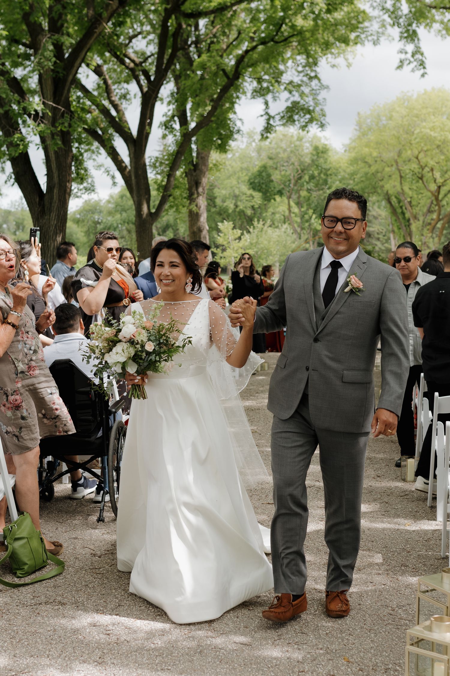 A joyful wedding couple walks down an outdoor aisle surrounded by guests under shady green trees.