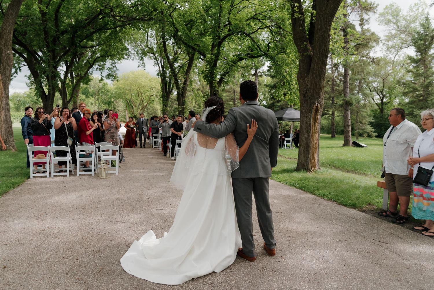 A couple stands together at an outdoor wedding ceremony with guests seated along a tree-lined path.