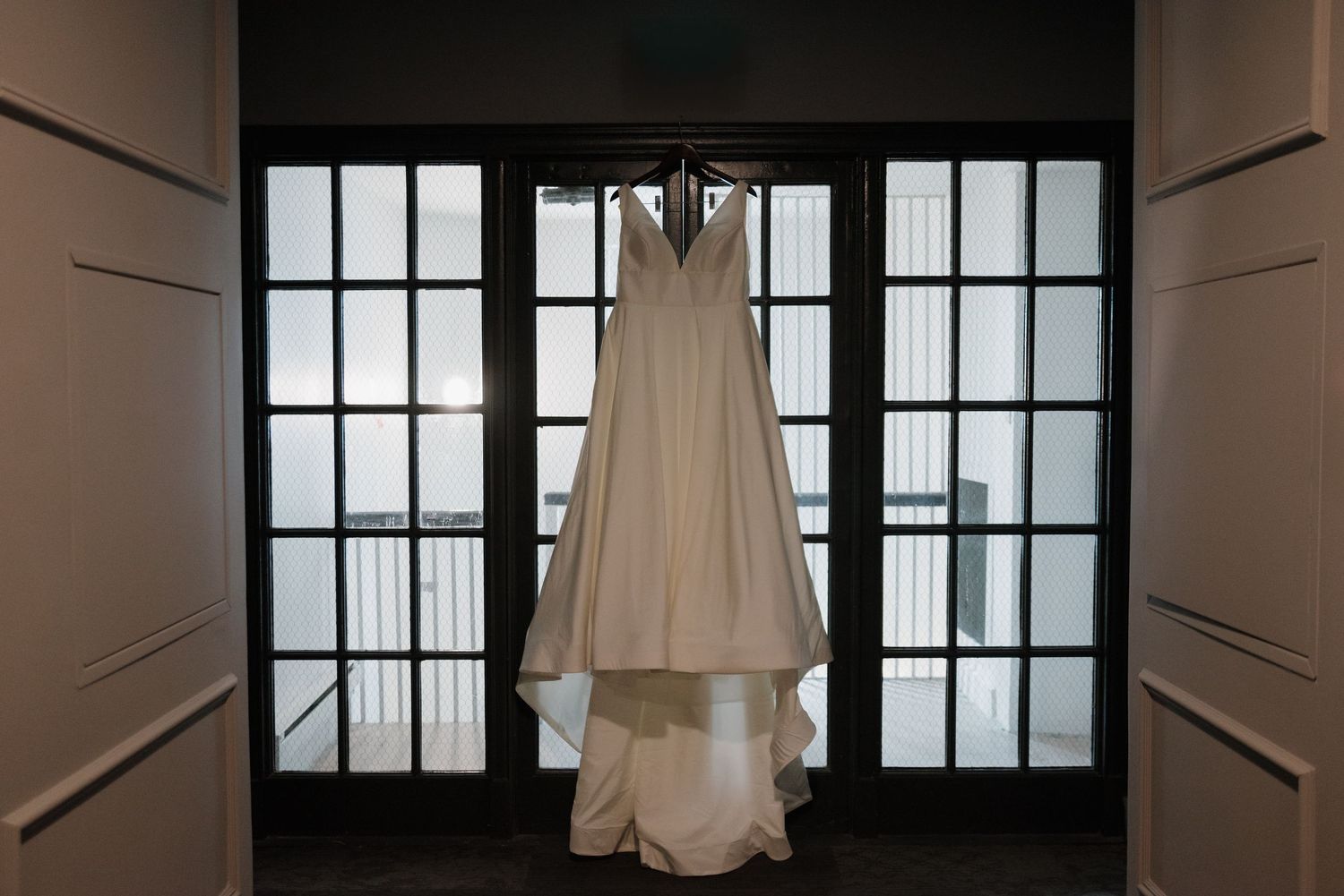 A white wedding dress hangs elegantly against glass paneled doors in a dimly lit room.