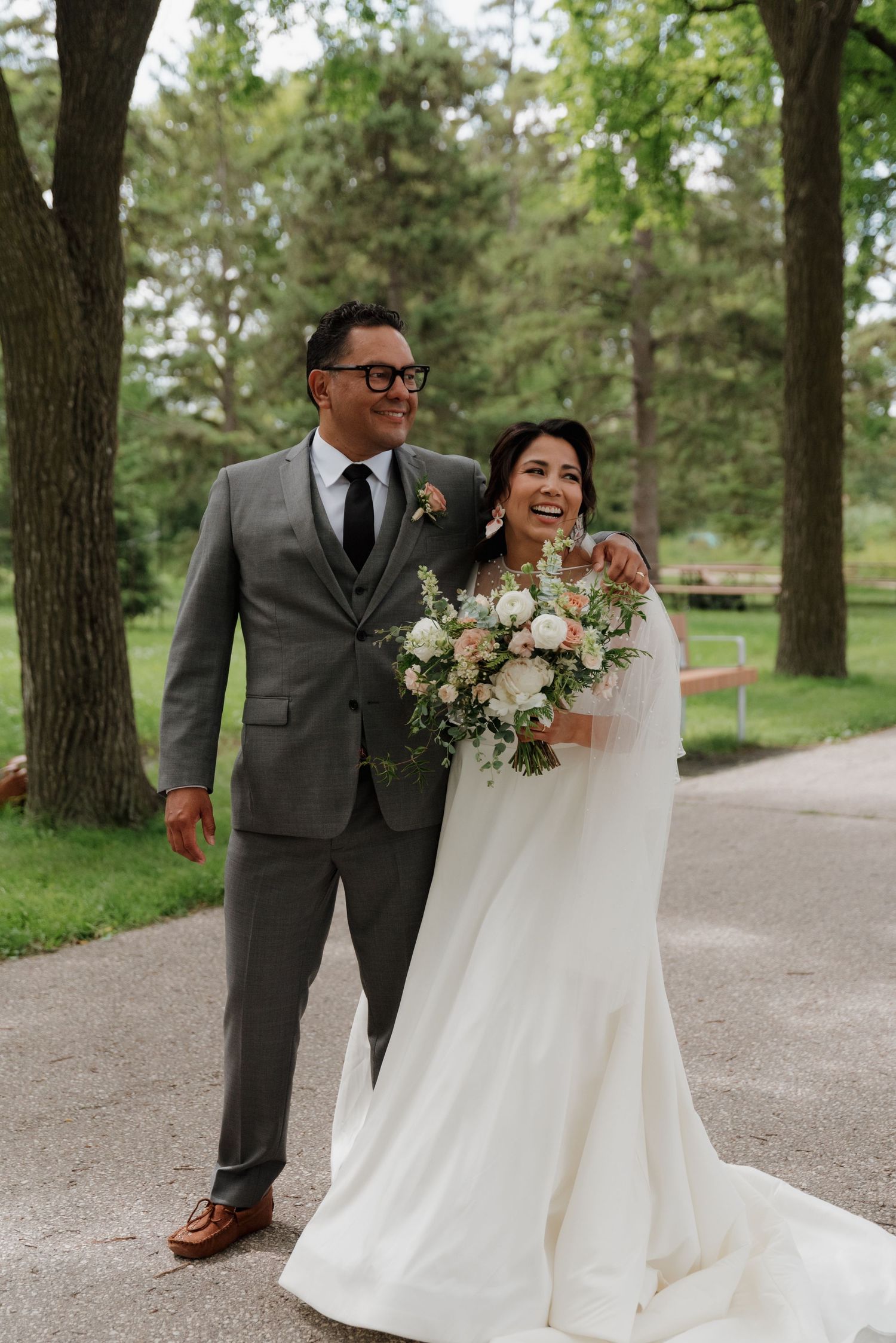 Happy couple in wedding attire laughing and walking together along a tree-lined path outdoors.