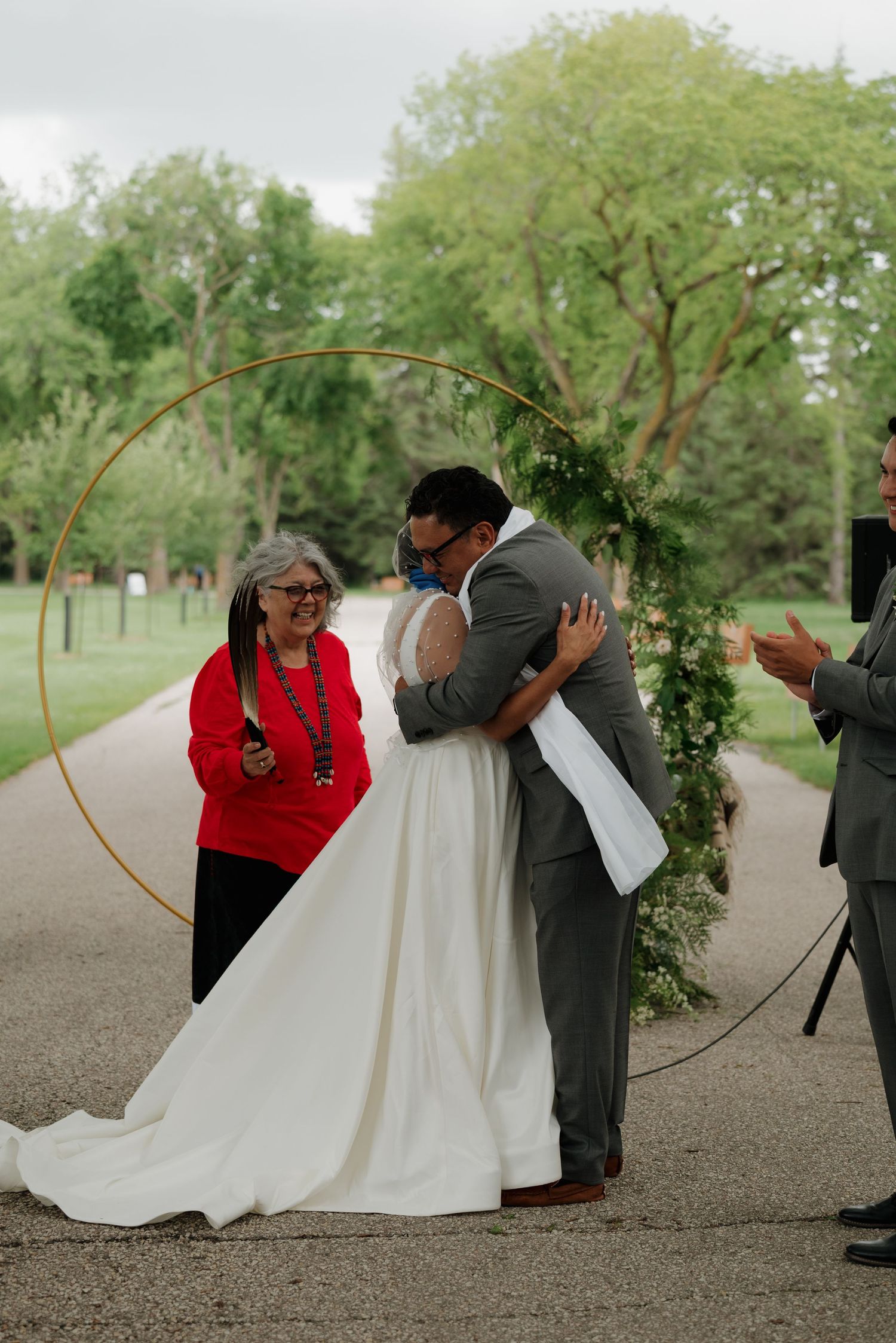 A couple shares their wedding ceremony outdoors in front of a curved floral arch with a person in red officiating.