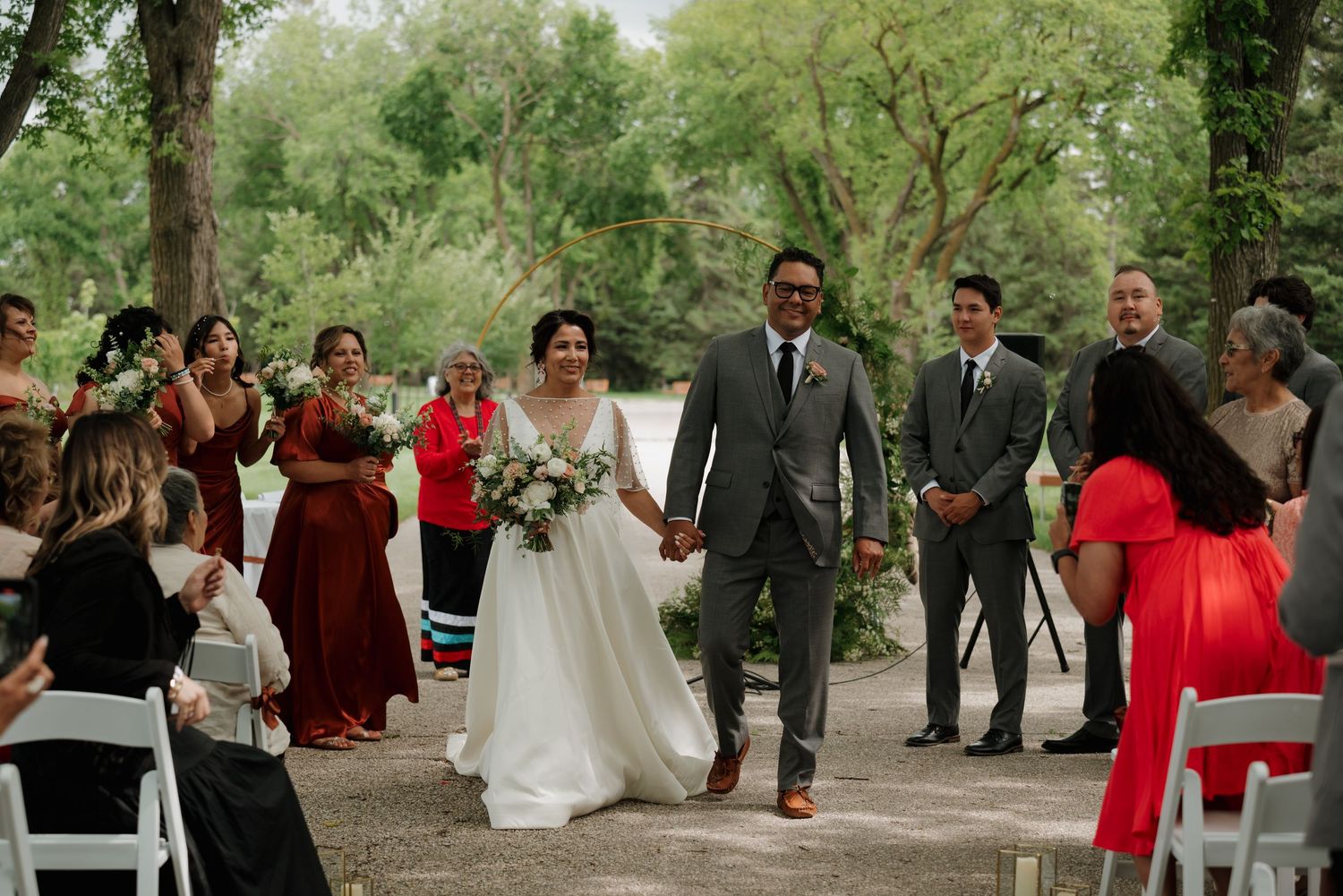 Newlyweds walk down outdoor aisle while wedding guests clap and celebrate at nature-filled ceremony.