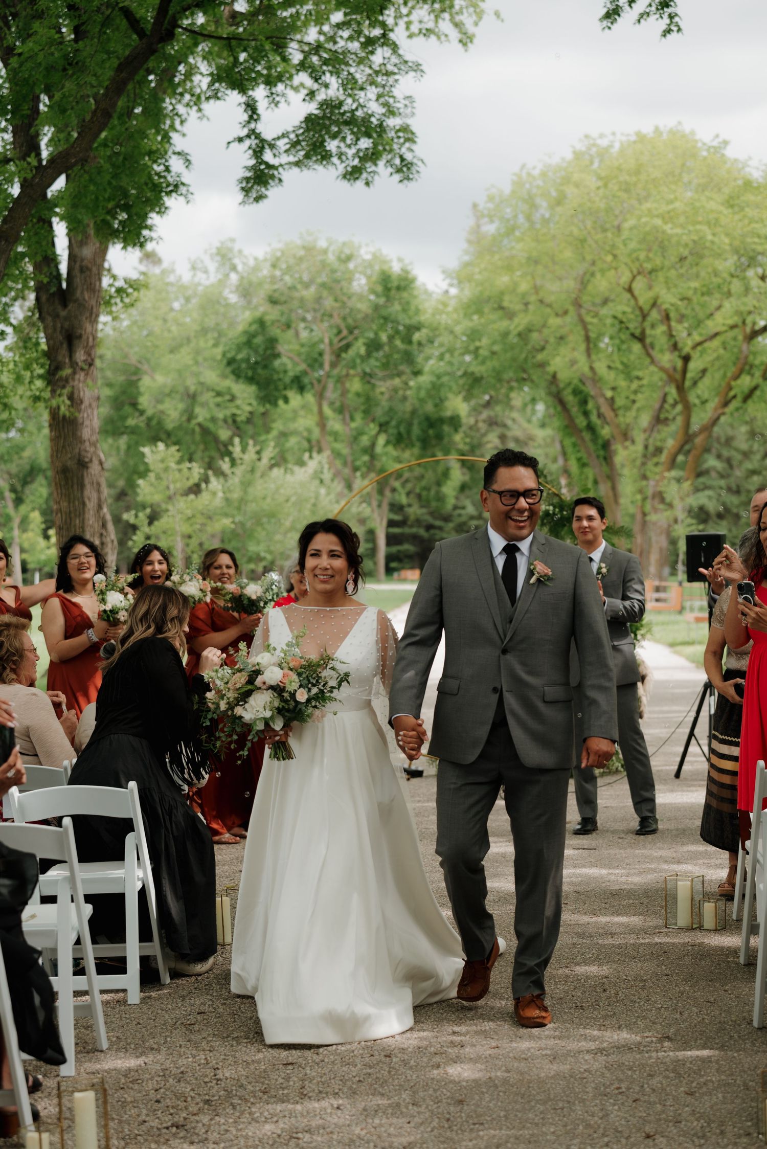 A wedding couple walks down an outdoor aisle lined with white chairs and trees while guests cheer and celebrate.