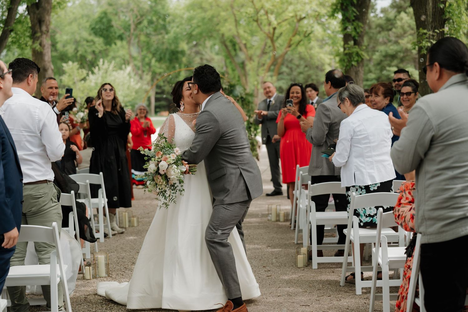 A couple dances during an outdoor wedding ceremony surrounded by white chairs and guests beneath shady trees.