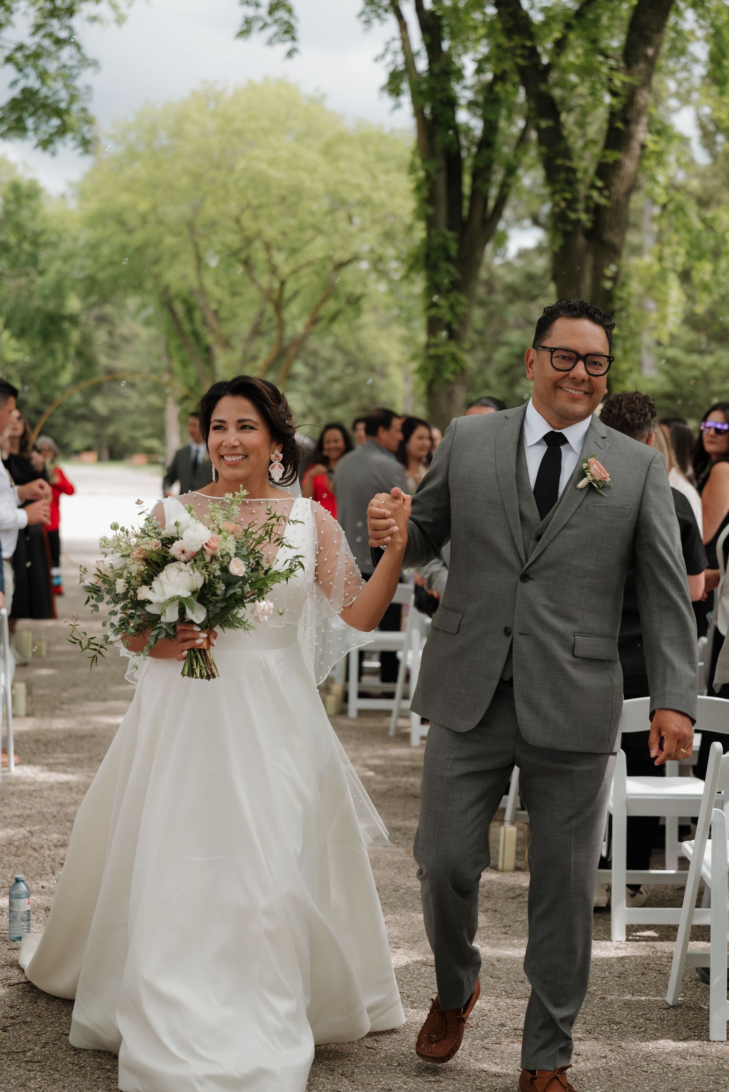 Newlyweds walk hand in hand down an aisle surrounded by guests and lush green trees.