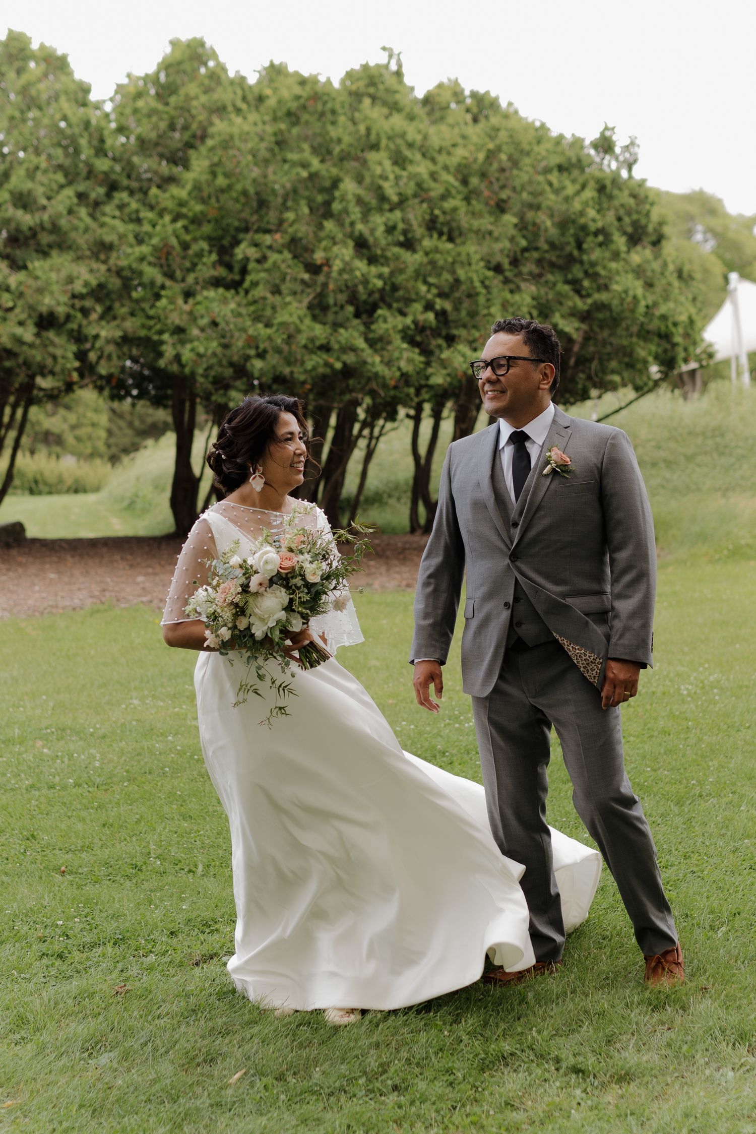 A newlywed couple walks together on a grassy hill with trees in the background, bride holding a white floral bouquet.
