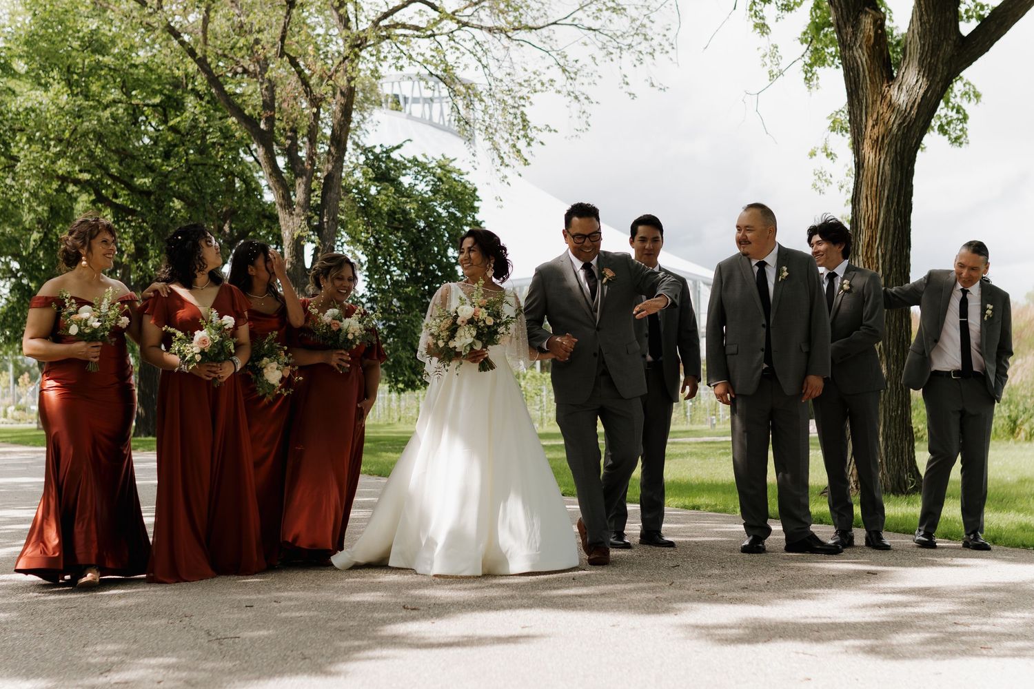 A wedding party stands together on a paved pathway with bridesmaids in burgundy dresses and groomsmen in dark suits.
