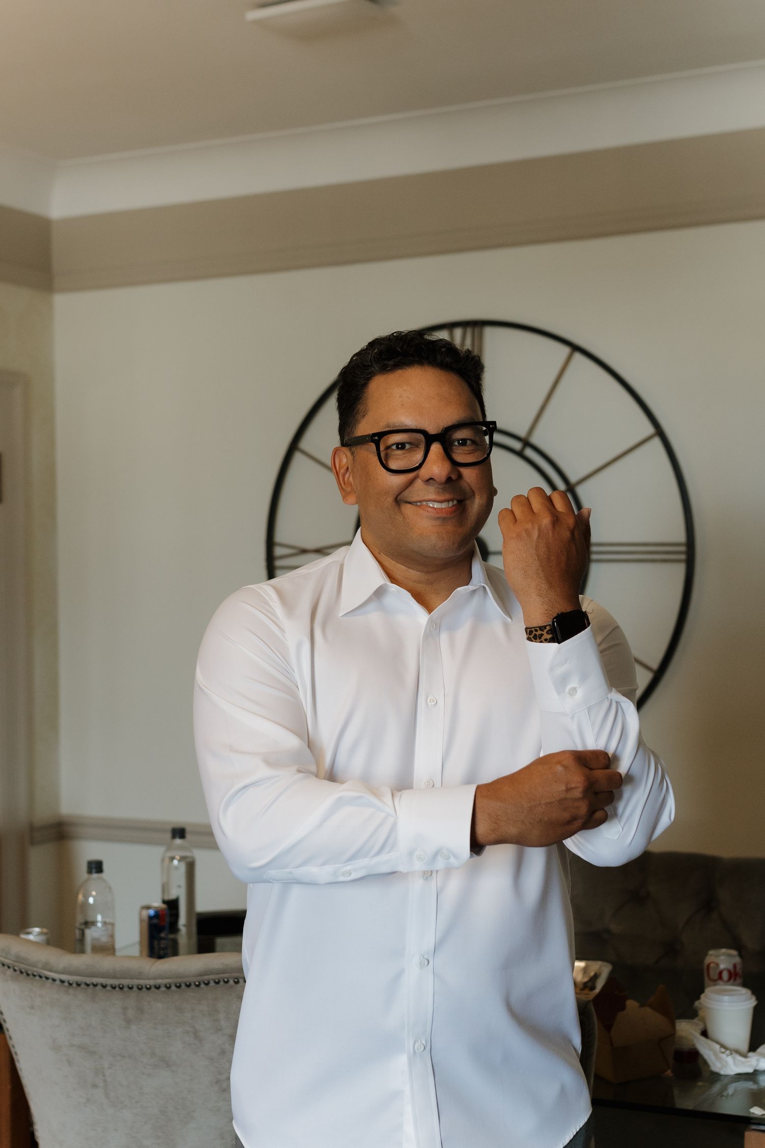 Person in white dress shirt and glasses adjusting cufflinks beside wall clock.