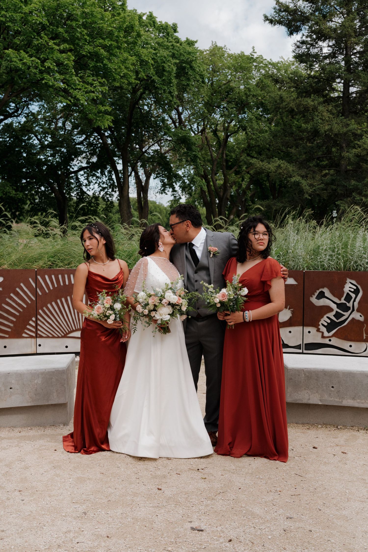 A wedding party in burgundy dresses poses together outdoors near modern architectural elements.