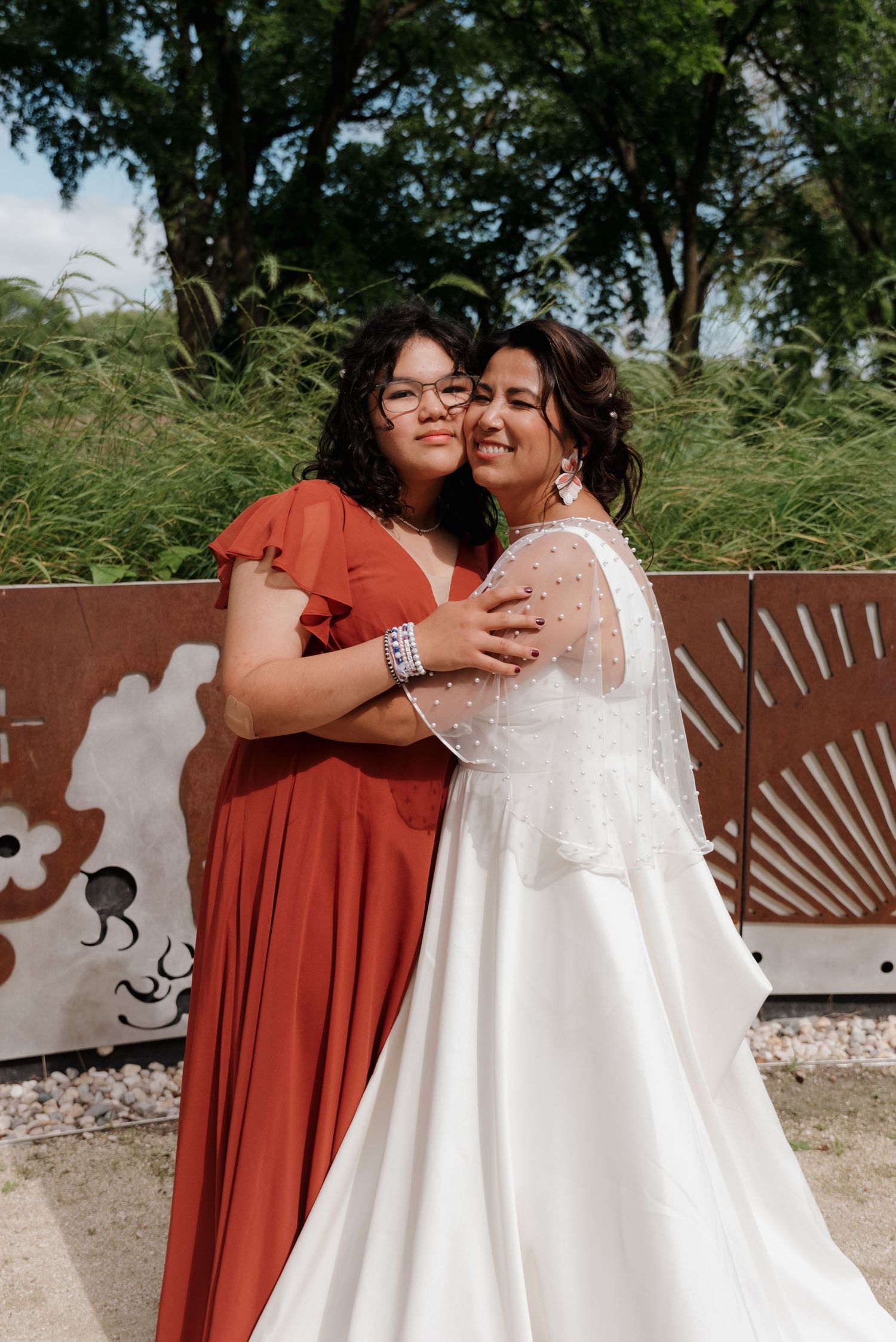 A bride in a white gown embraces another person in a rust-colored dress against a decorative outdoor backdrop.