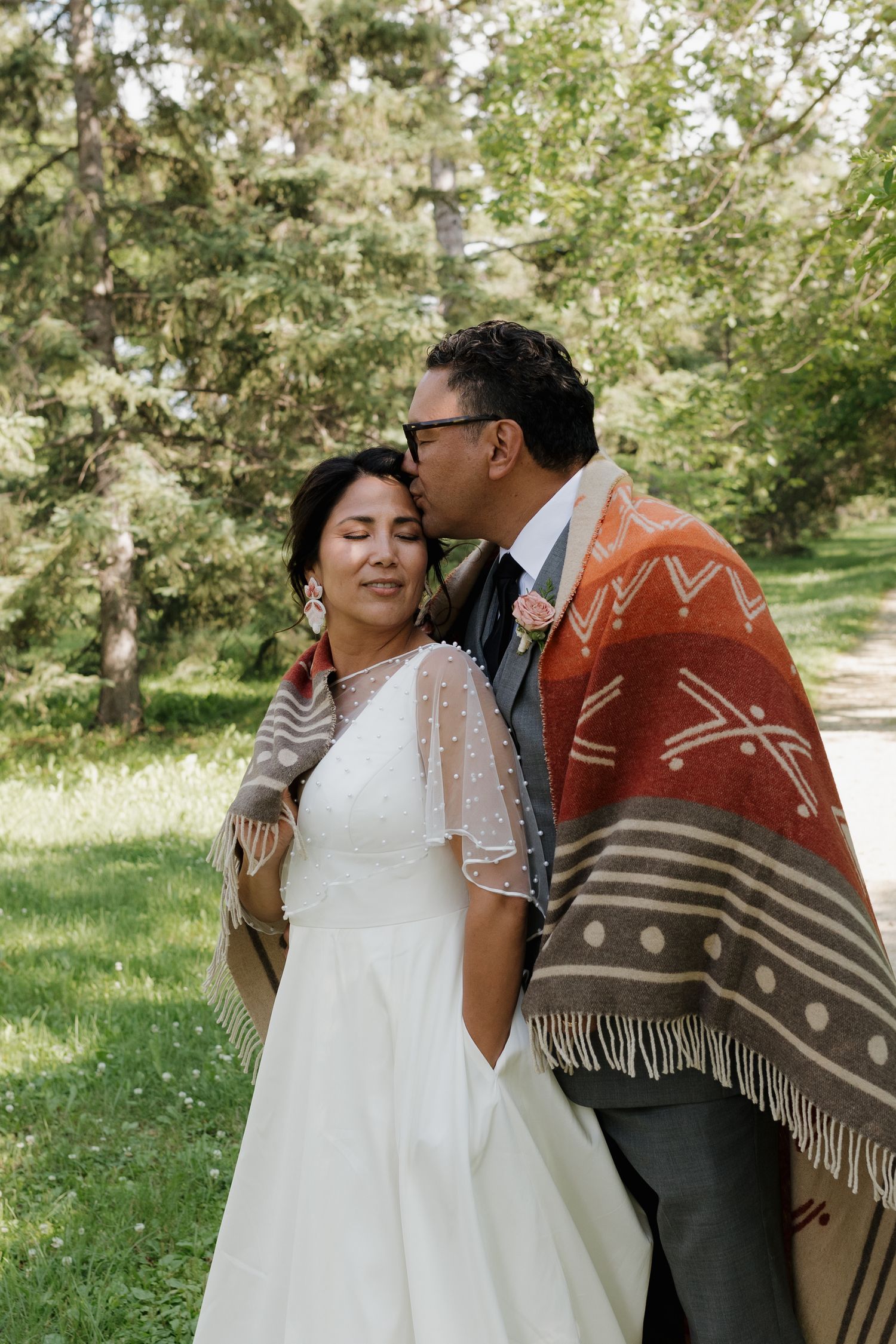 A couple shares an intimate moment wrapped in a decorative blanket with geometric patterns in a garden setting.