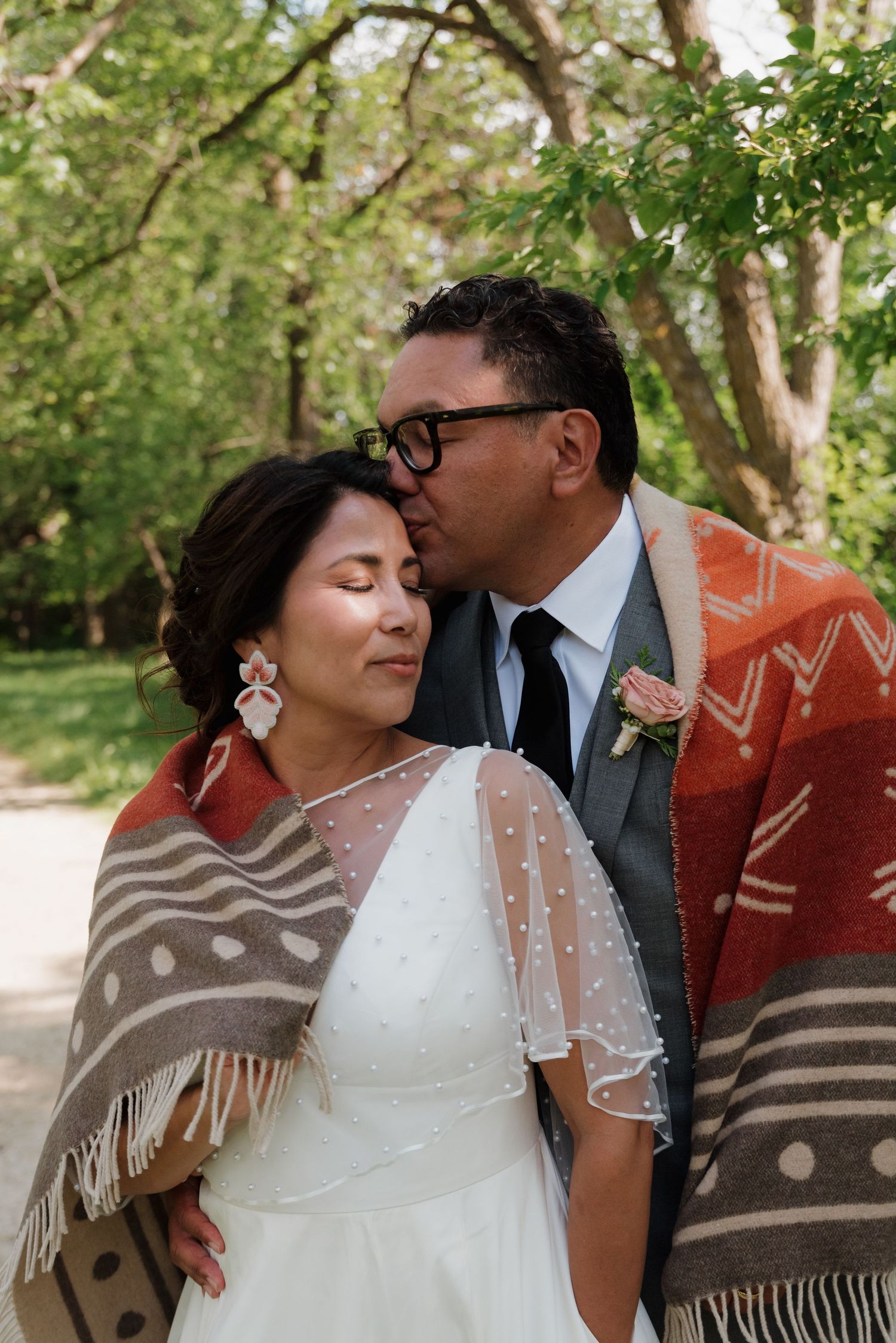Two people share an intimate moment outdoors wrapped in patterned blankets with trees in the background.
