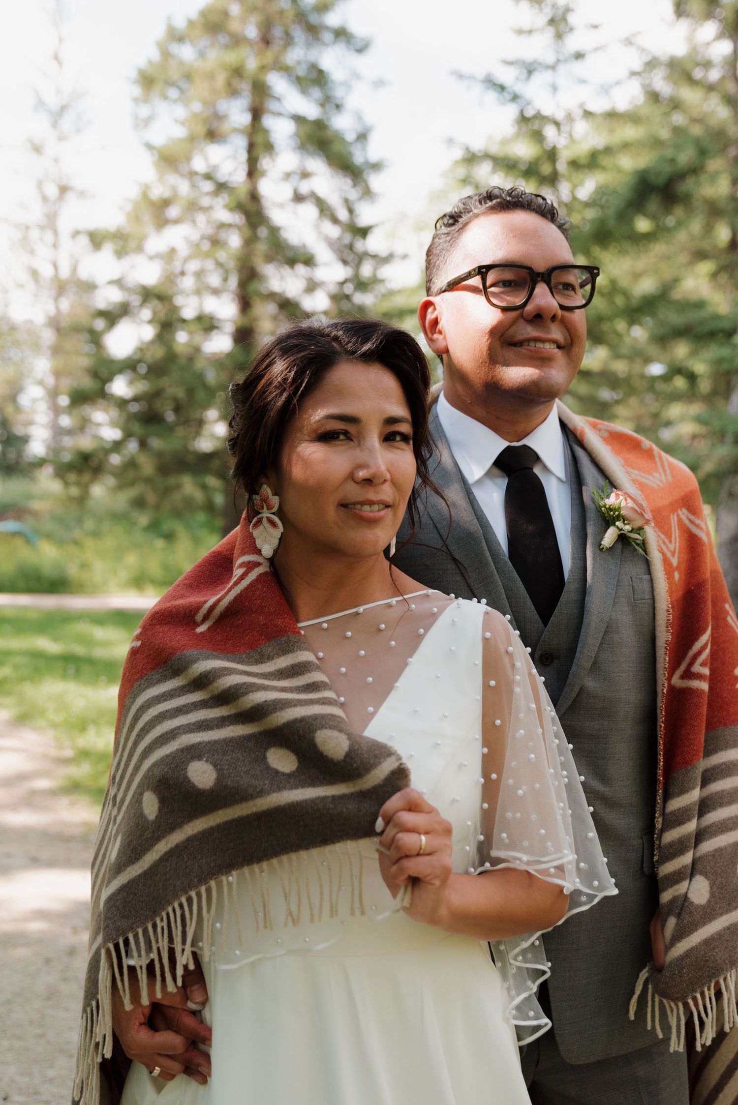 A newlywed couple poses outdoors wearing a traditional wedding dress and grey suit with a patterned blanket wrap.