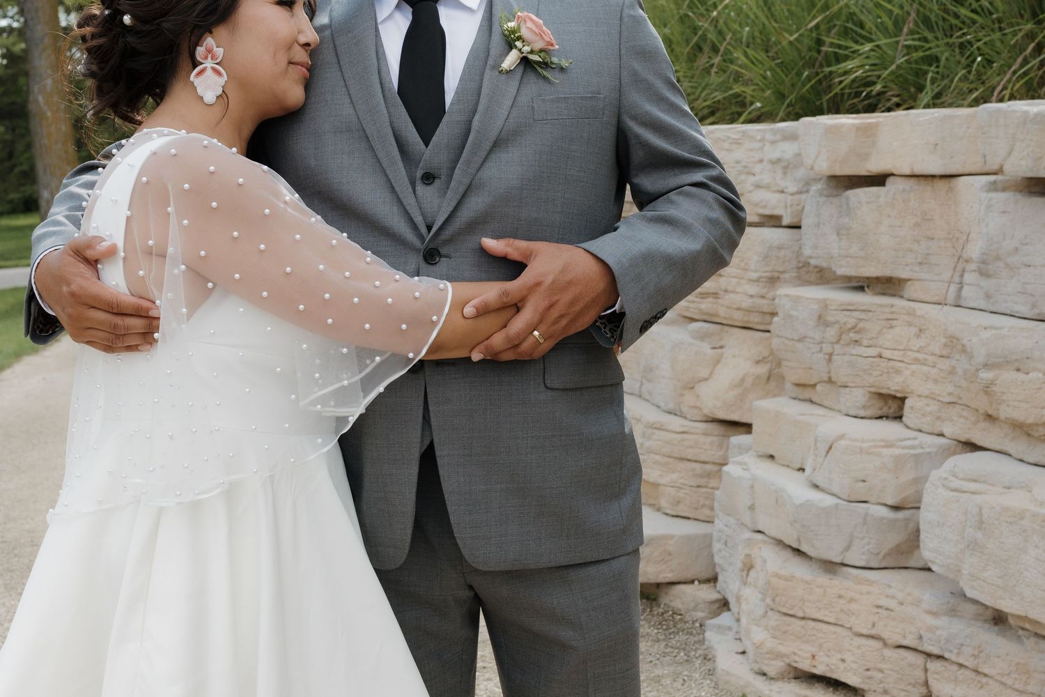 An elegant wedding couple embraces near a stone wall, with the bride wearing a sheer-sleeved white gown and groom in grey suit.