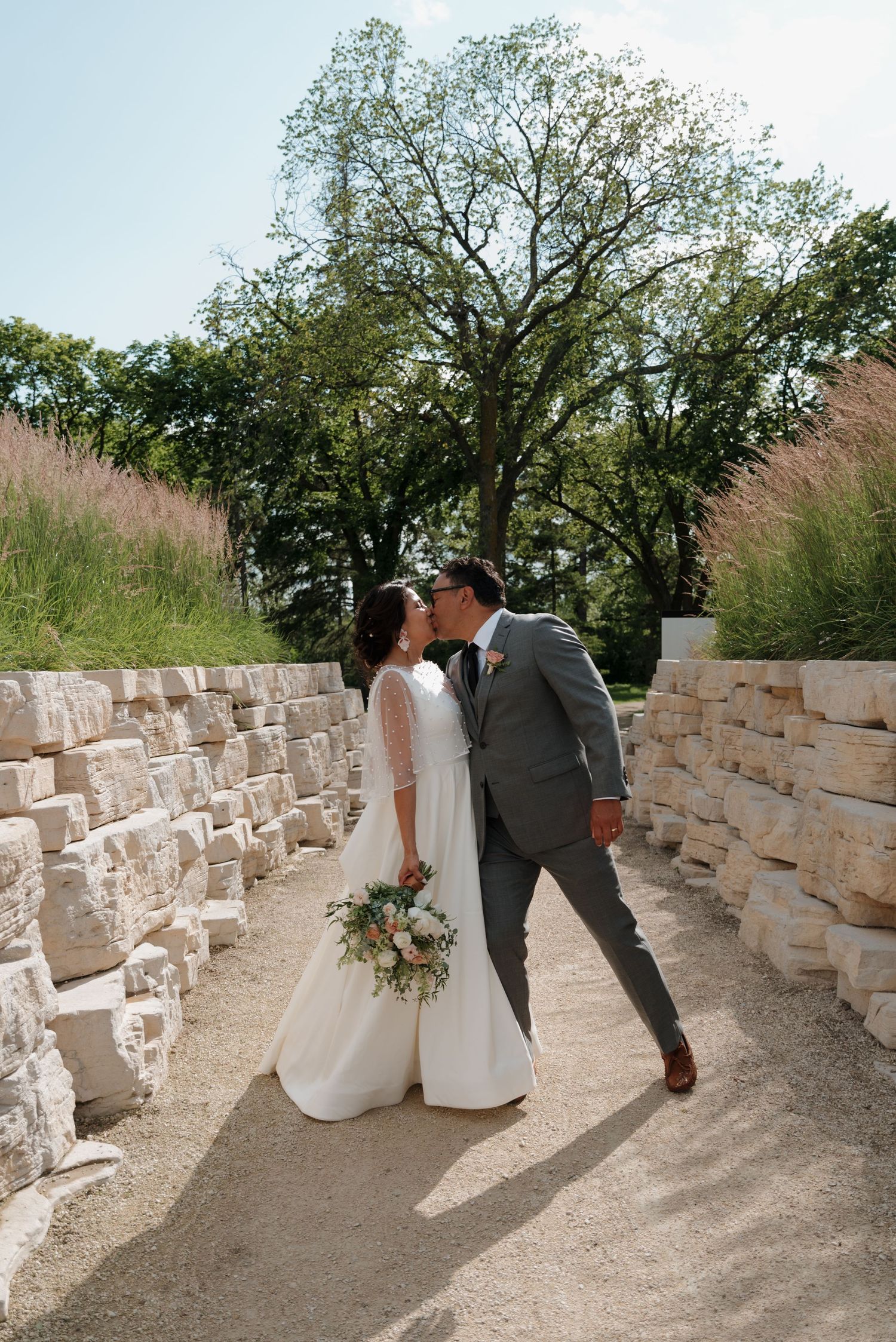 A romantic wedding photo of a couple between stone walls with trees in the background.