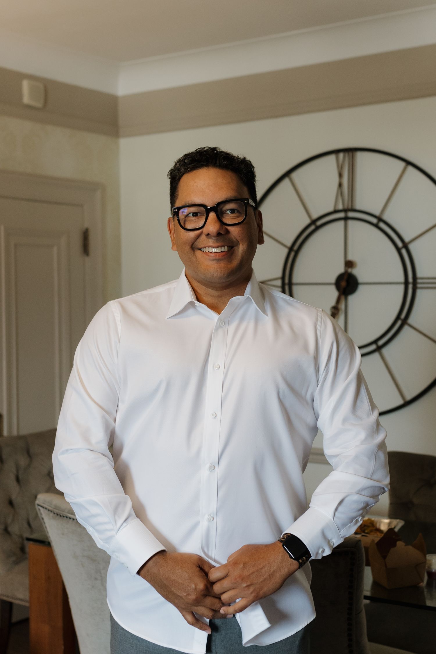 A person in a white dress shirt and glasses stands smiling in front of a large wall clock.