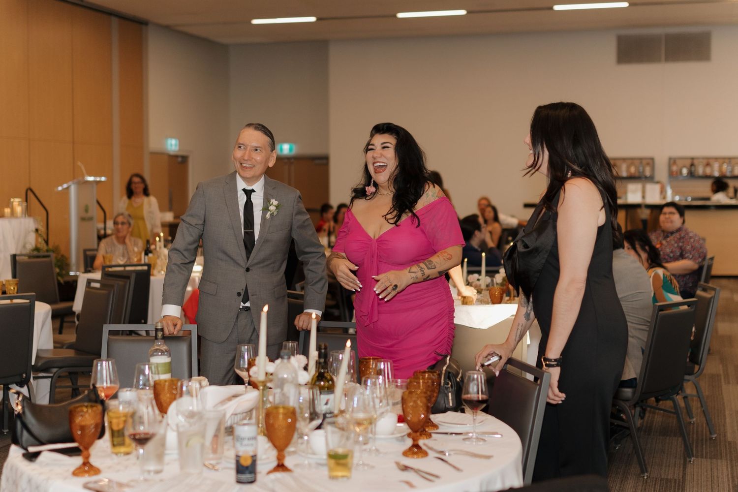 Wedding reception guests gather around an elegant table with amber-colored glasses and formal place settings.