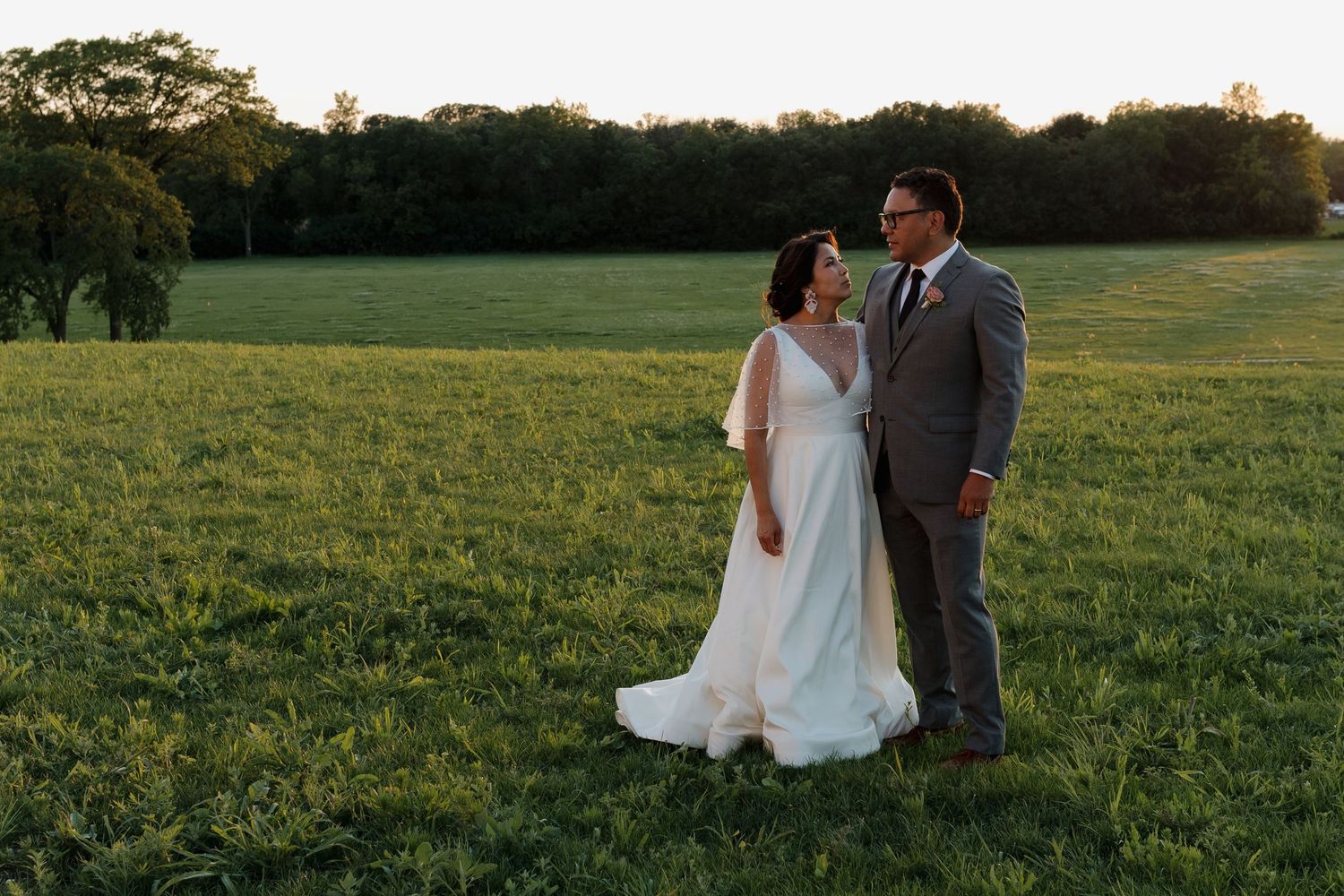 Newlyweds pose together in a grassy field during golden hour.