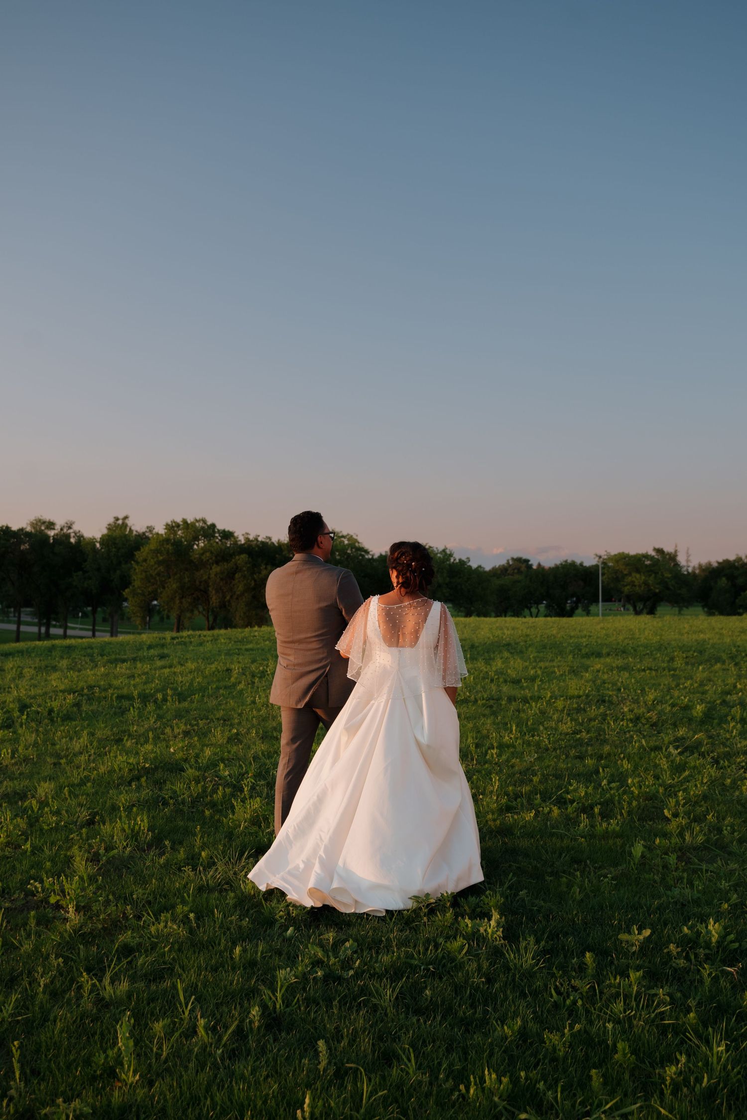 A couple in wedding attire standing together on a grassy field during golden hour sunset.