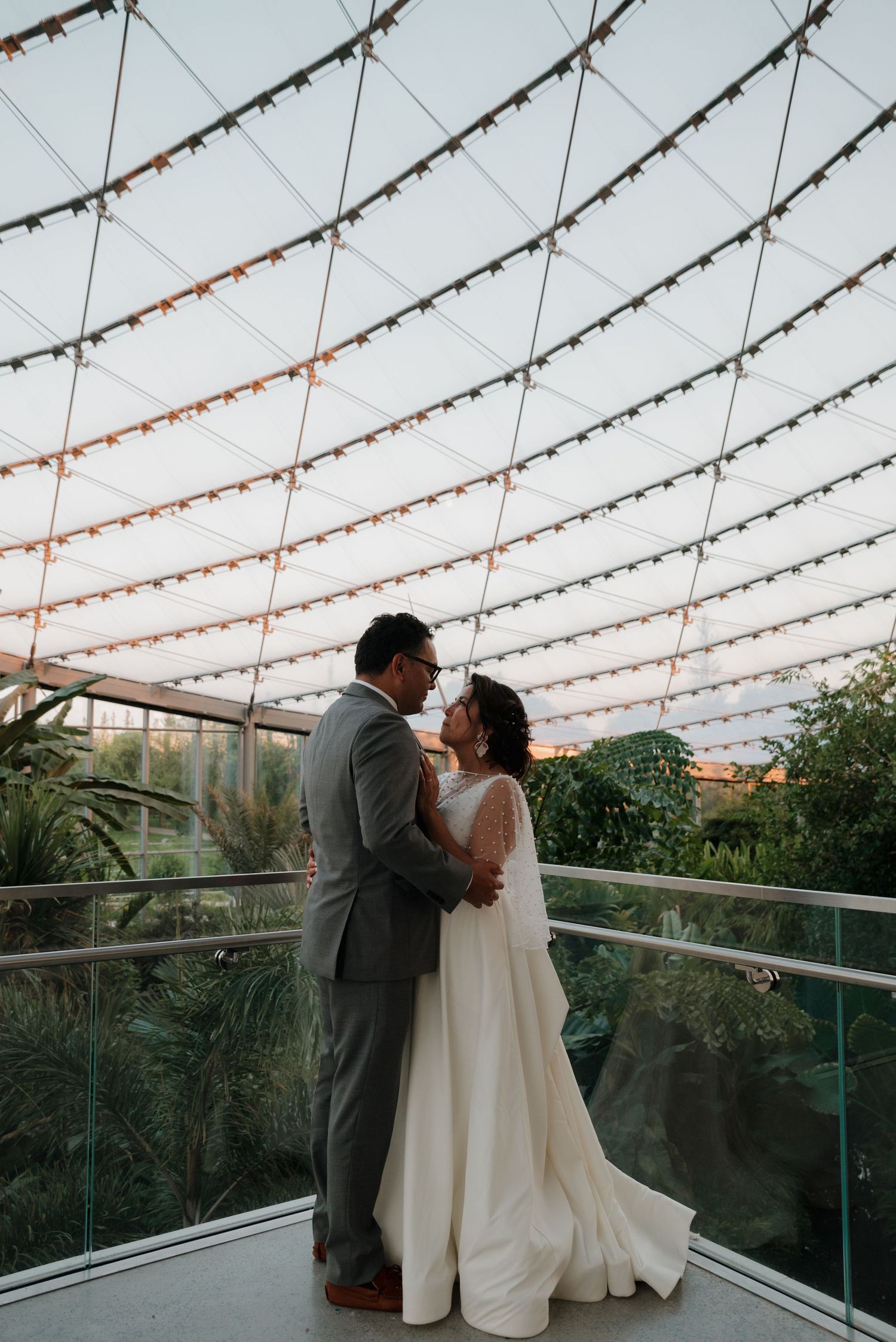 A couple shares a romantic moment under string lights on a balcony overlooking lush greenery at sunset.