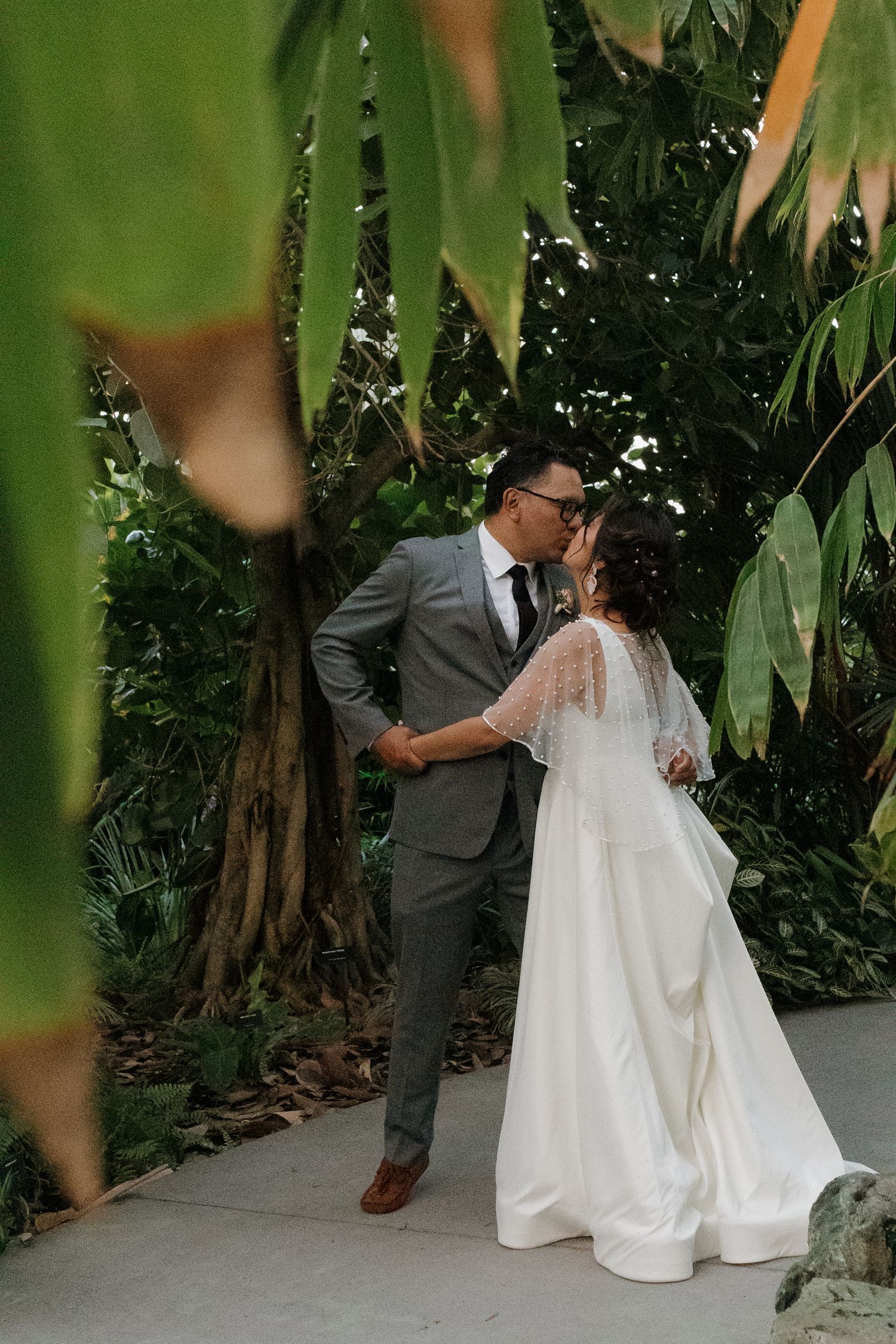 A romantic wedding couple poses among tropical foliage, with the bride in white and groom in gray suit.
