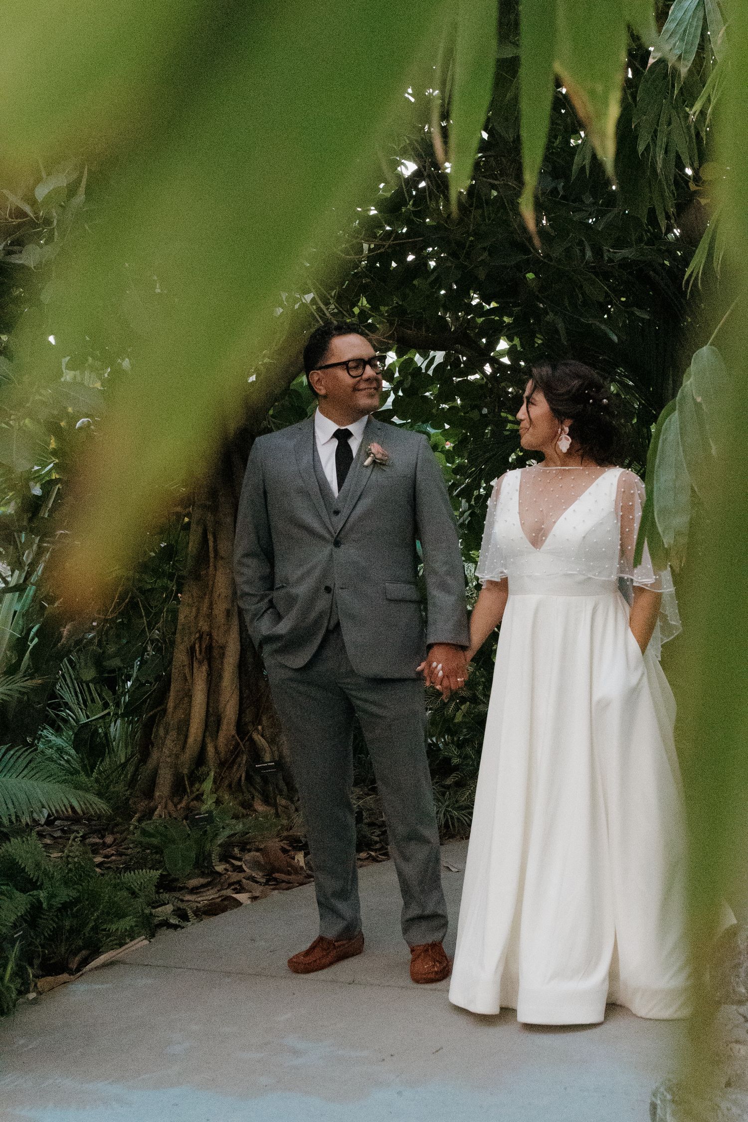 A couple in wedding attire holds hands while walking along a tropical garden path with lush greenery.