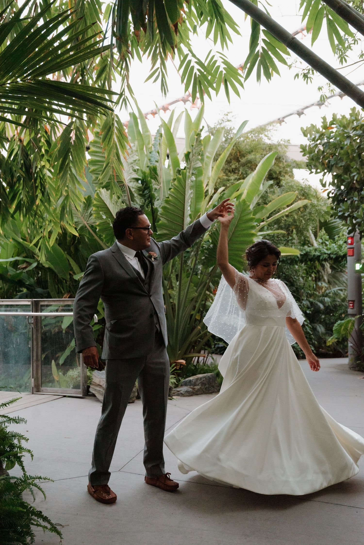 A newlywed couple dances joyfully in a garden setting with palm trees and tropical foliage.