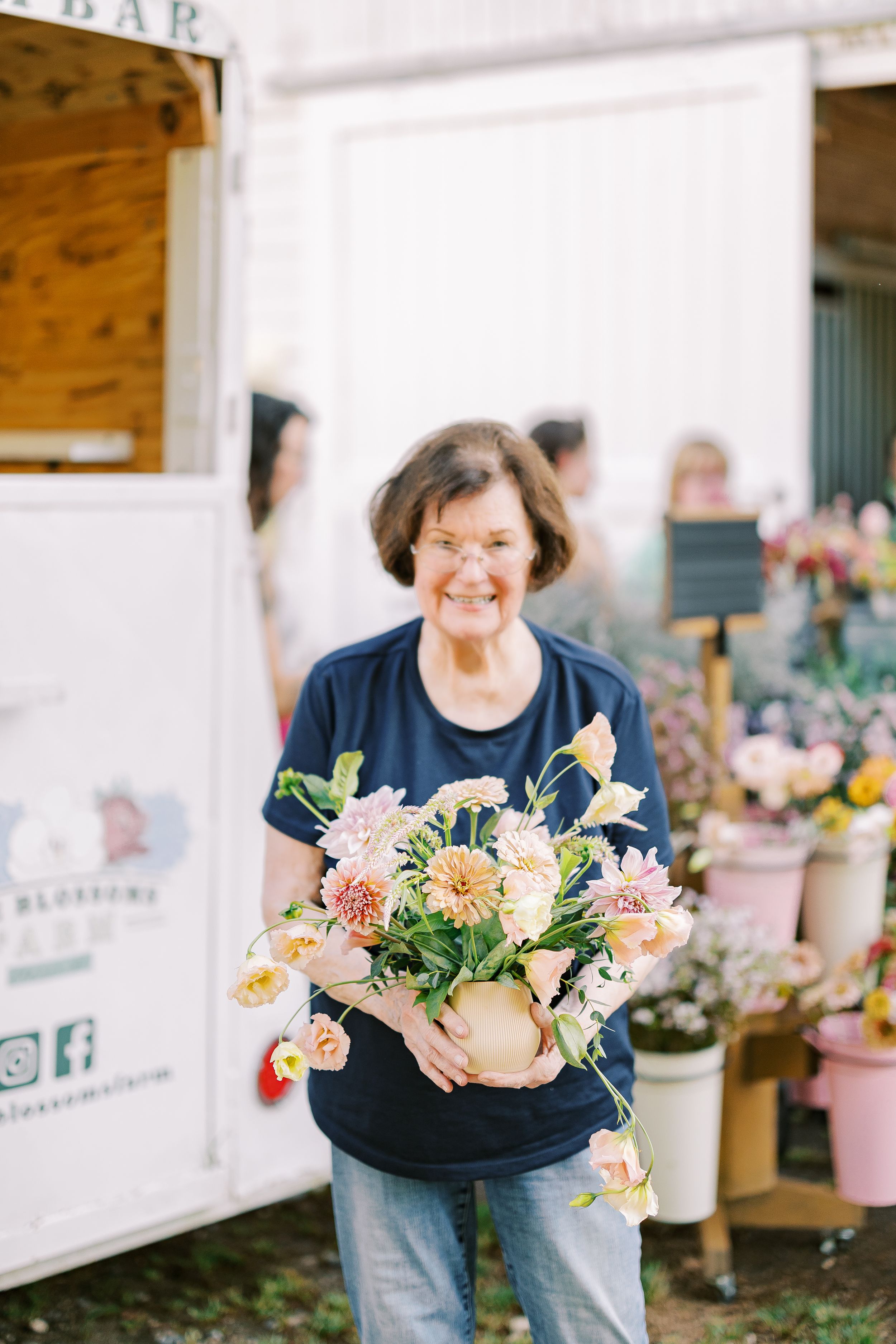 Person in denim and navy holding beautiful peach and pink floral arrangements at an outdoor market.